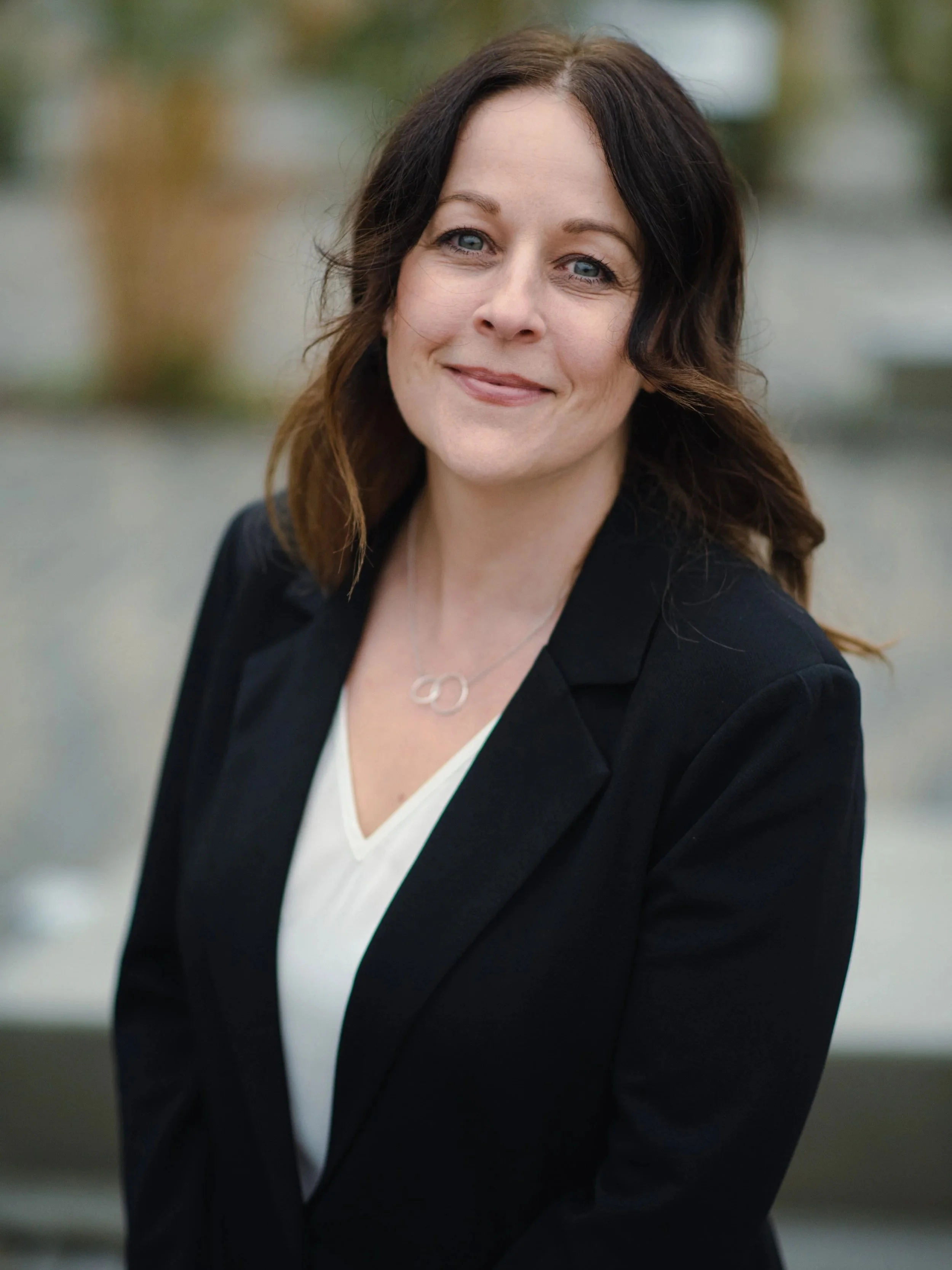 A woman with shoulder-length brown hair and blue eyes smiling outdoors, wearing a black blazer and a white top, with a blurred background.