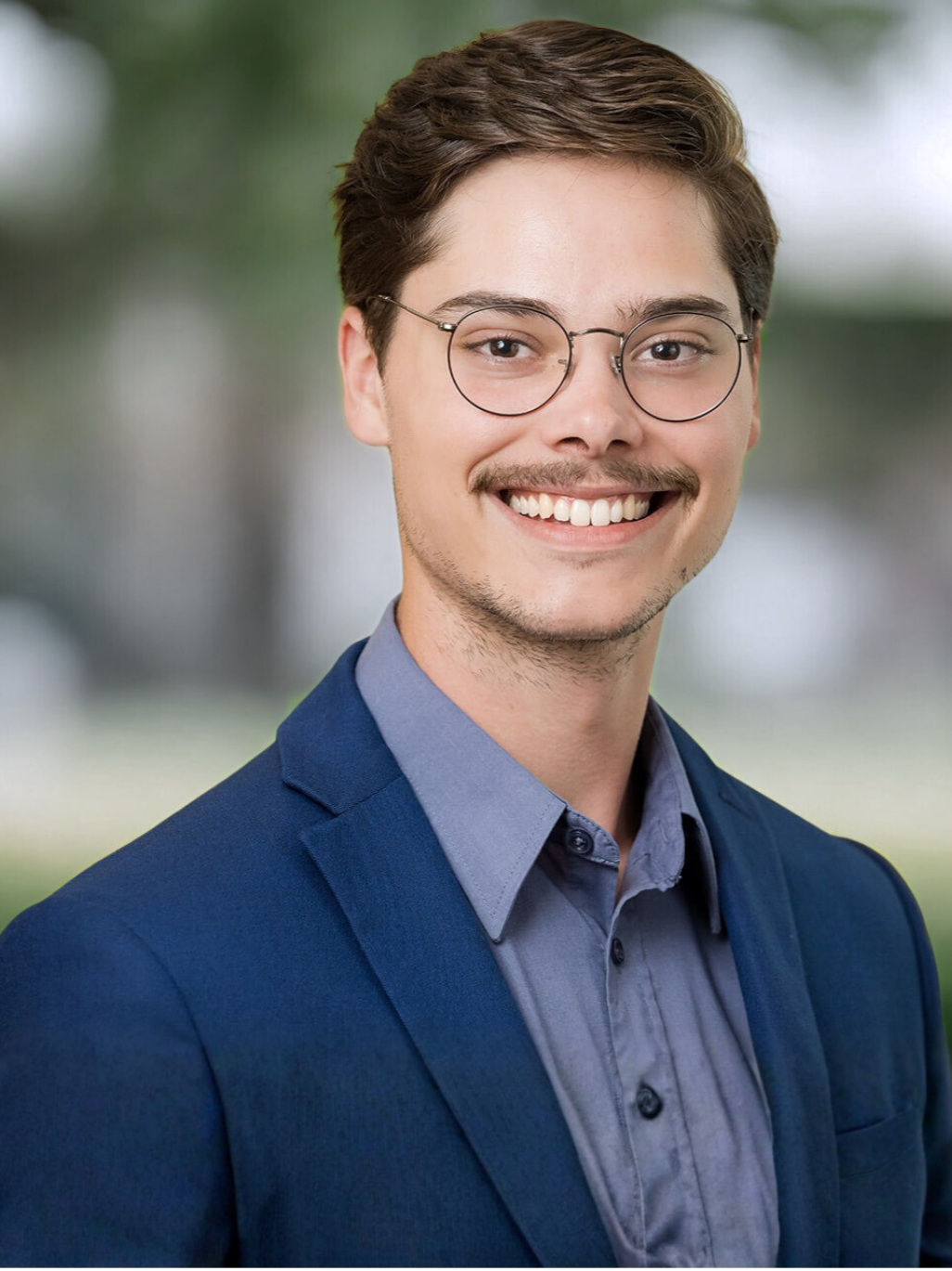 Smiling young man with short brown hair, glasses, and a mustache, wearing a blue suit jacket and a gray shirt, standing outdoors with a blurred green background.