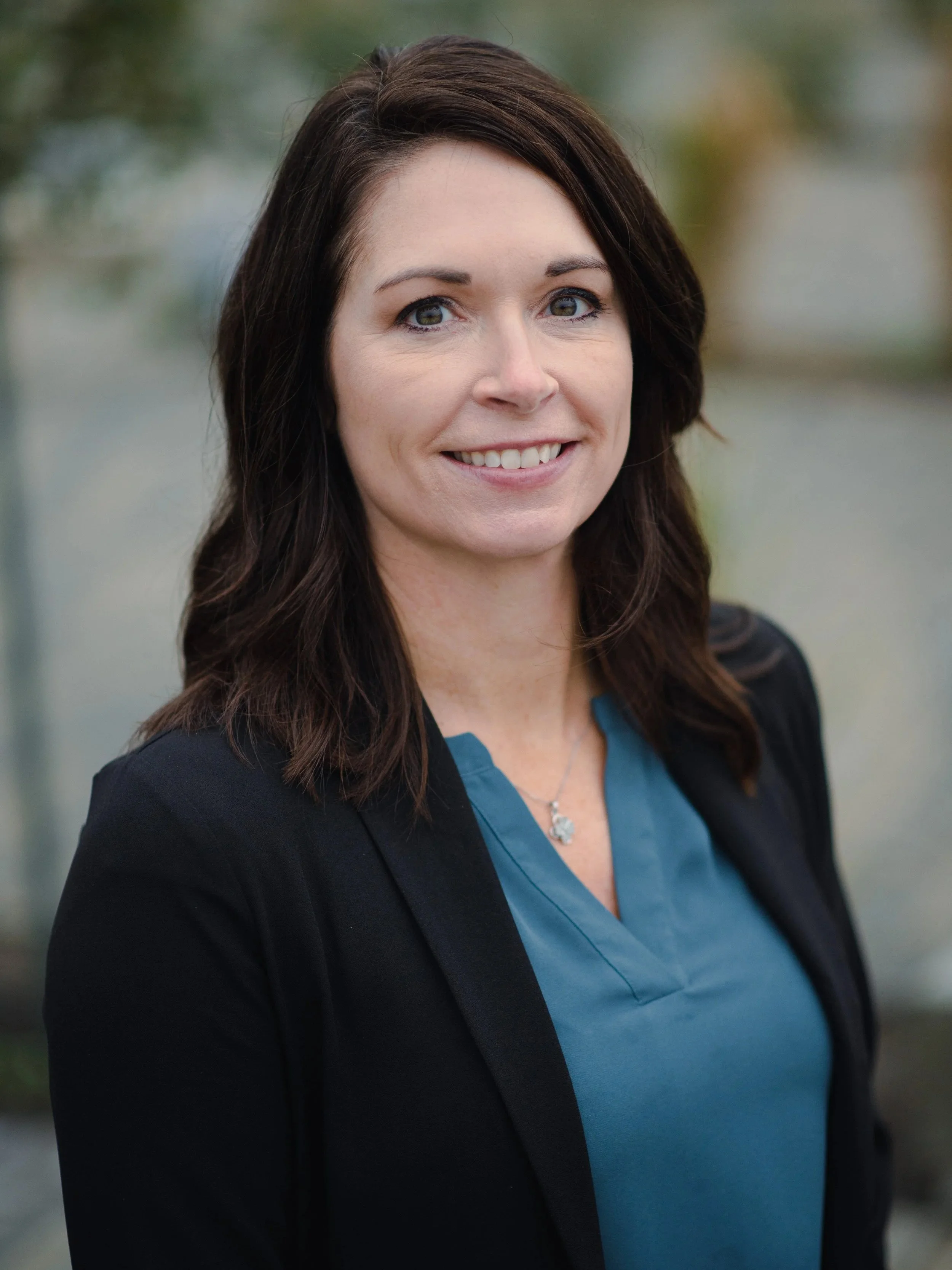 A woman with shoulder-length dark brown hair wearing a black blazer over a blue blouse, smiling outdoors with a blurred natural background.