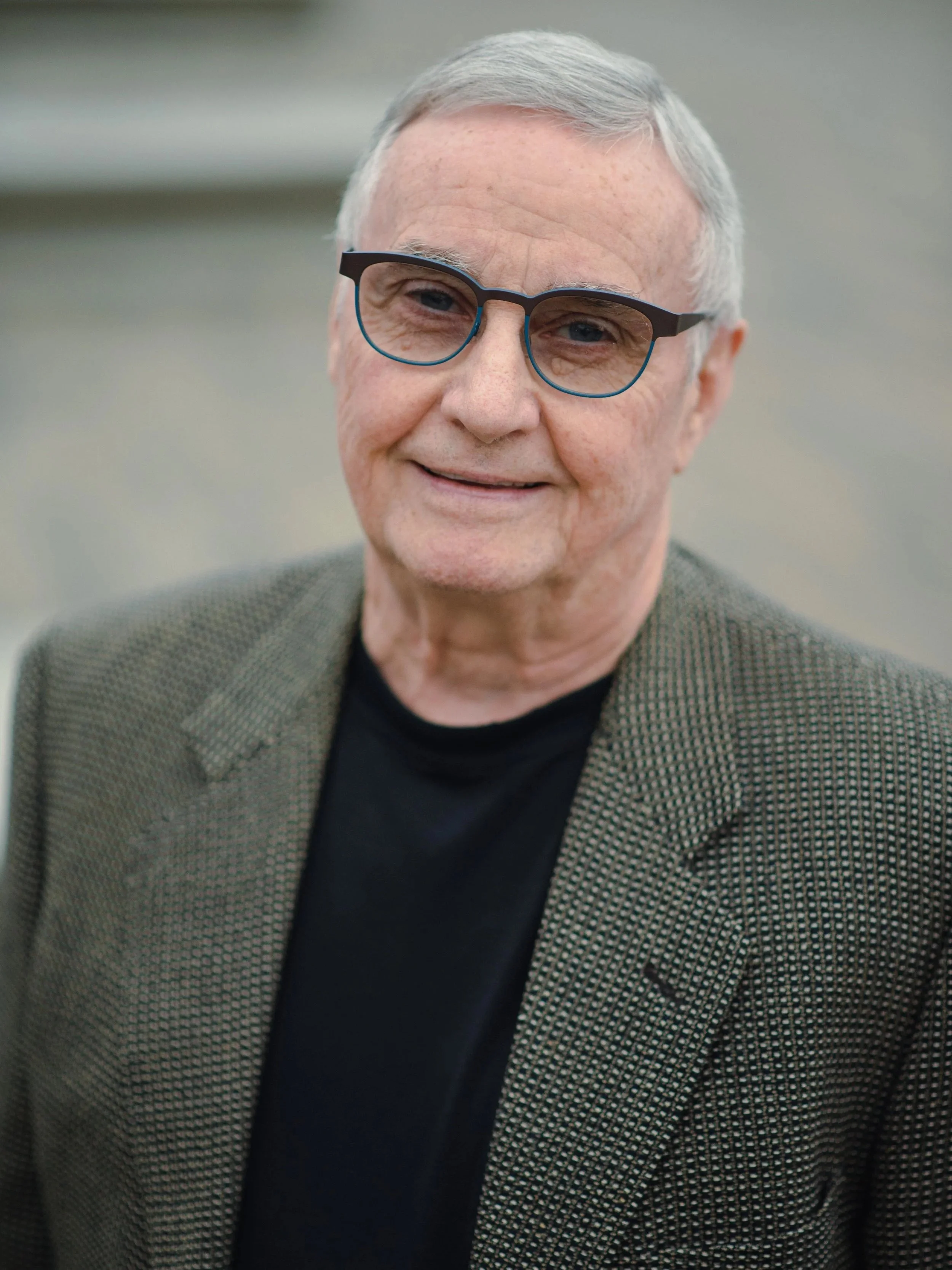 Photograph of an elderly man with gray hair wearing black-rimmed glasses, a black shirt, and a brown checkered blazer, smiling at the camera.