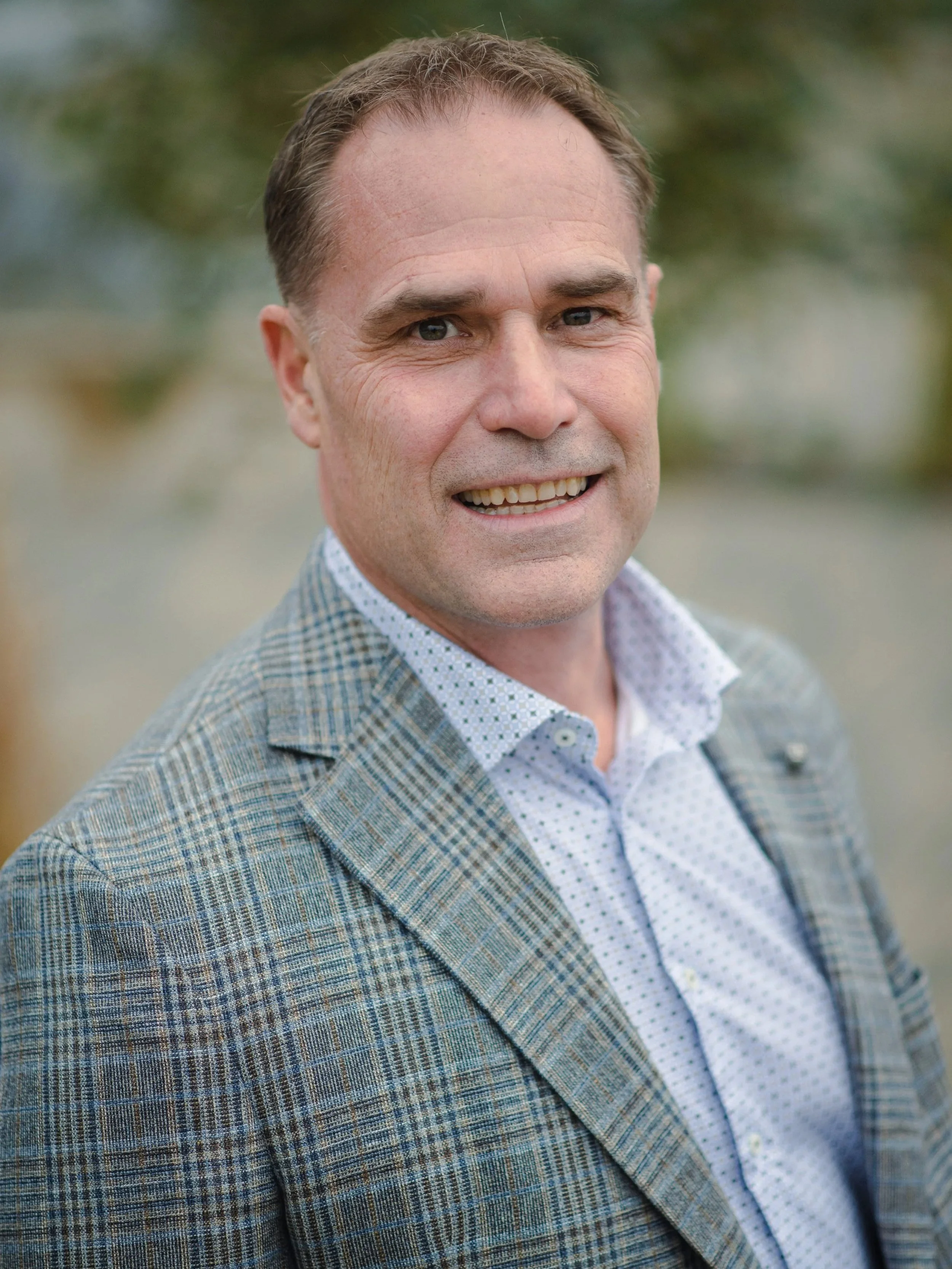 A middle-aged man with short brown hair and a friendly smile, wearing a checked blazer over a light patterned shirt, standing outdoors with a blurred green and beige background.