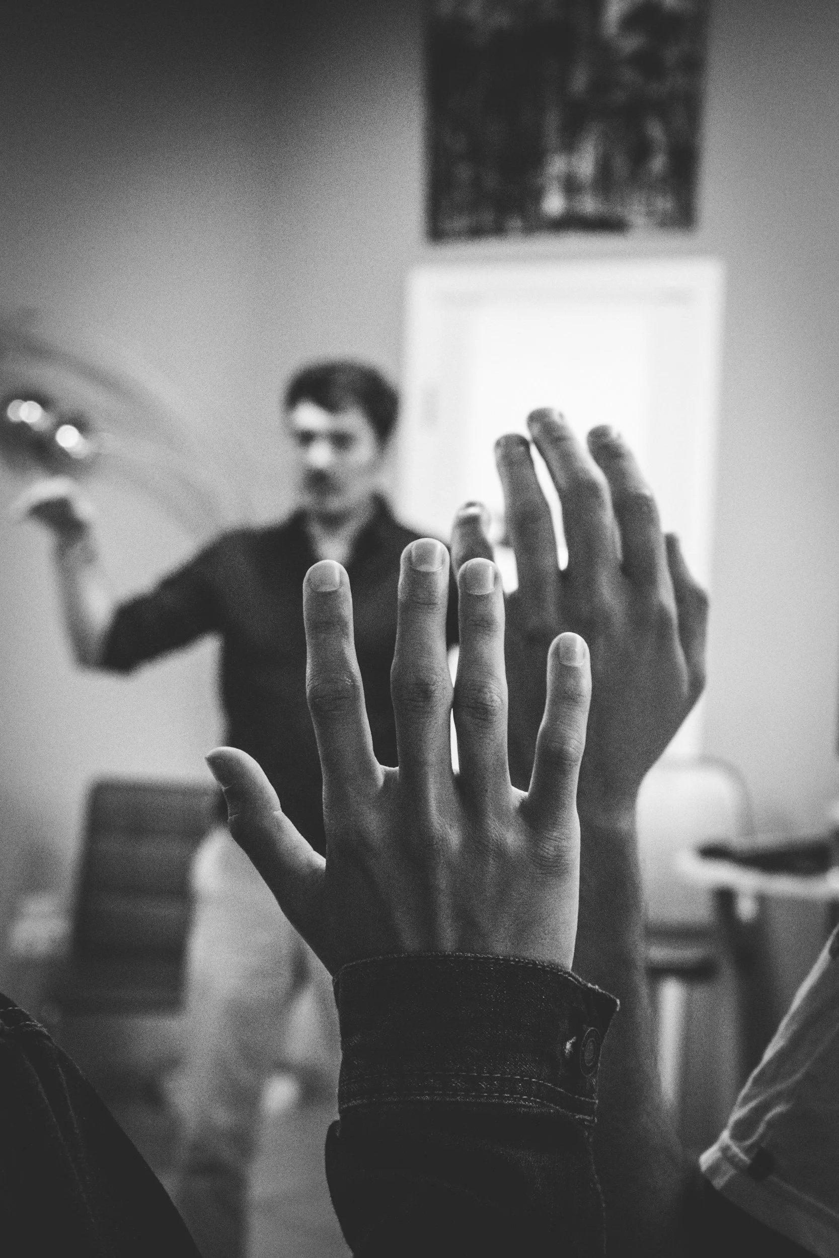 Two hands are raised in the foreground with fingers extended, and a person is blurred in the background gesturing with one hand, indoors, black and white photo.