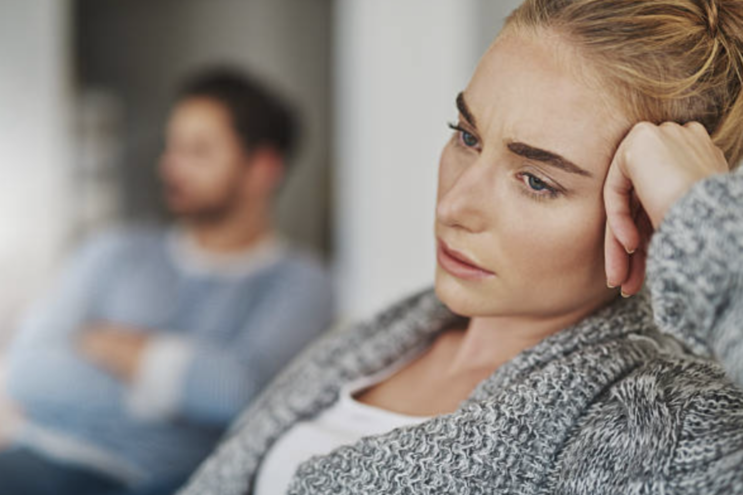 A woman with blonde hair resting her head on her hand, looking sad or worried, while a man in the background appears to be talking or drinking from a cup, blurred out.