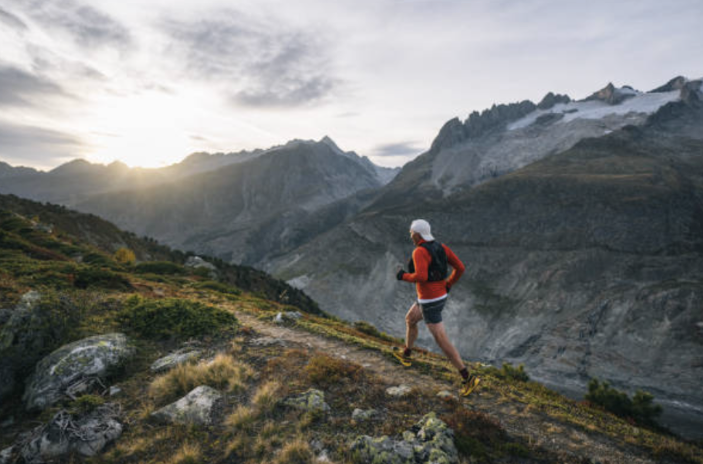 Person wearing red jacket, black shorts, and yellow running shoes trail running on a mountain trail with mountainous landscape and cloudy sky in the background at sunrise or sunset.