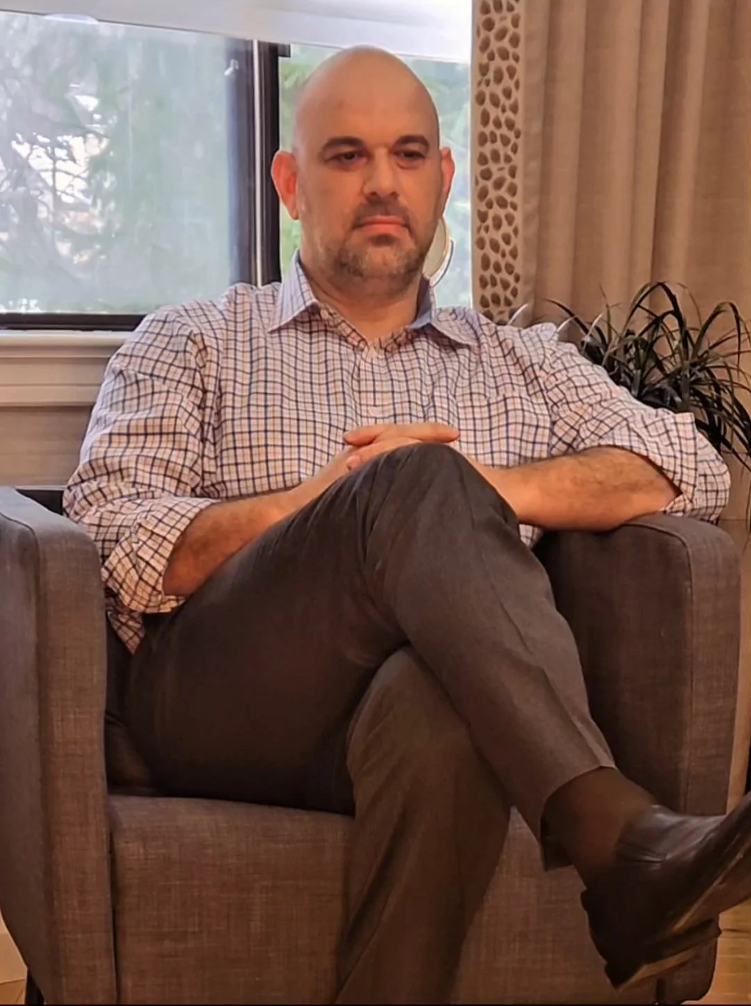 A bald man with a beard sitting in a chair with crossed legs, wearing a checkered shirt and dark pants, in a room with a window and curtains behind him.