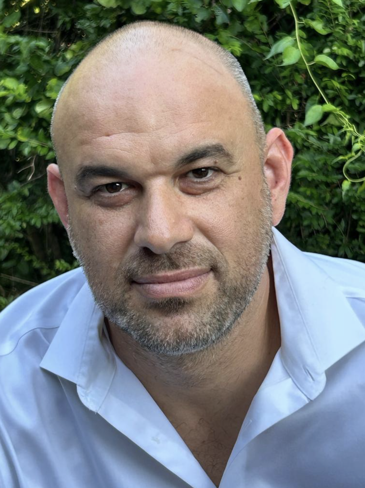 Close-up of a smiling man with a bald head and gray beard, wearing a light blue shirt, outdoors with green foliage in the background.