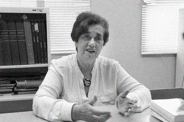 Mujer mayor sentada en una mesa, hablando y gesticulando, en una oficina con estantería y libros al fondo.