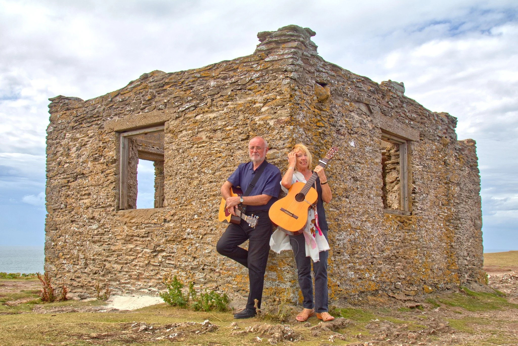 A man and woman standing with guitars in front of a weathered stone building with three window openings on a grassy landscape under a cloudy sky.