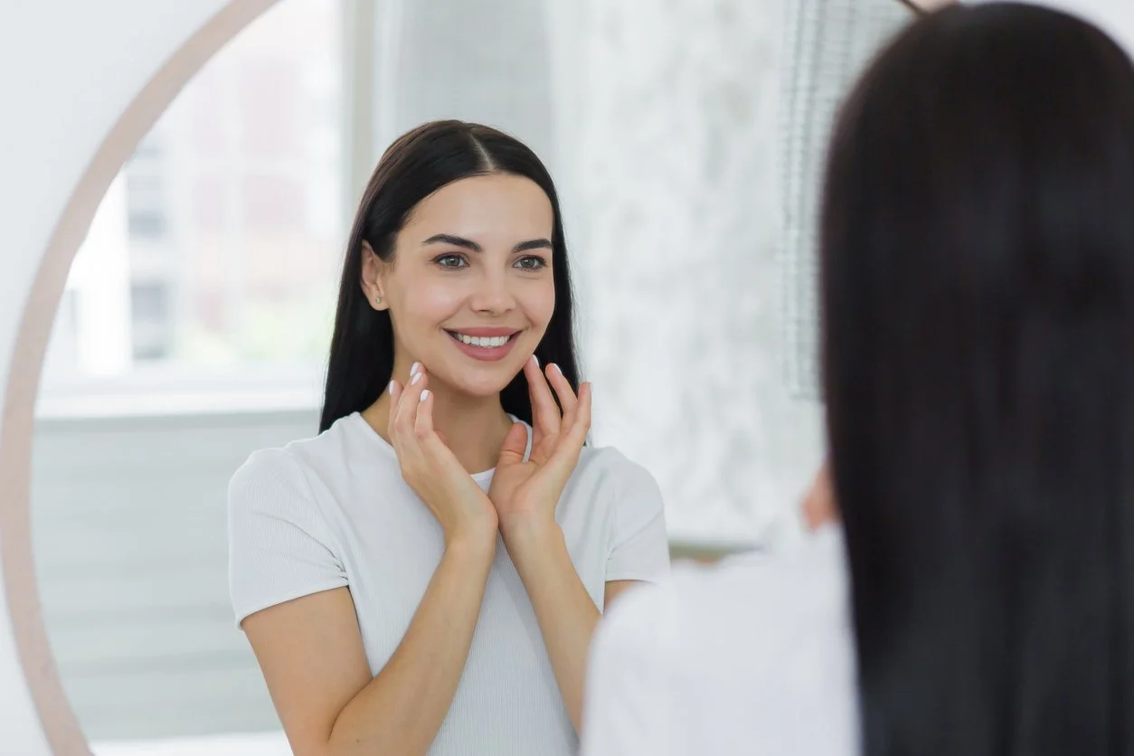 Young woman looking in the mirror at home, touching her face and smiling, admiring her face