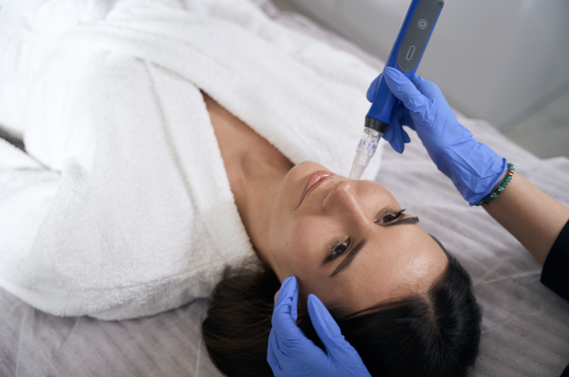 A woman lying on a bed in a robe receiving a cosmetic treatment with a handheld device, while a person wearing blue gloves holds her head steady.