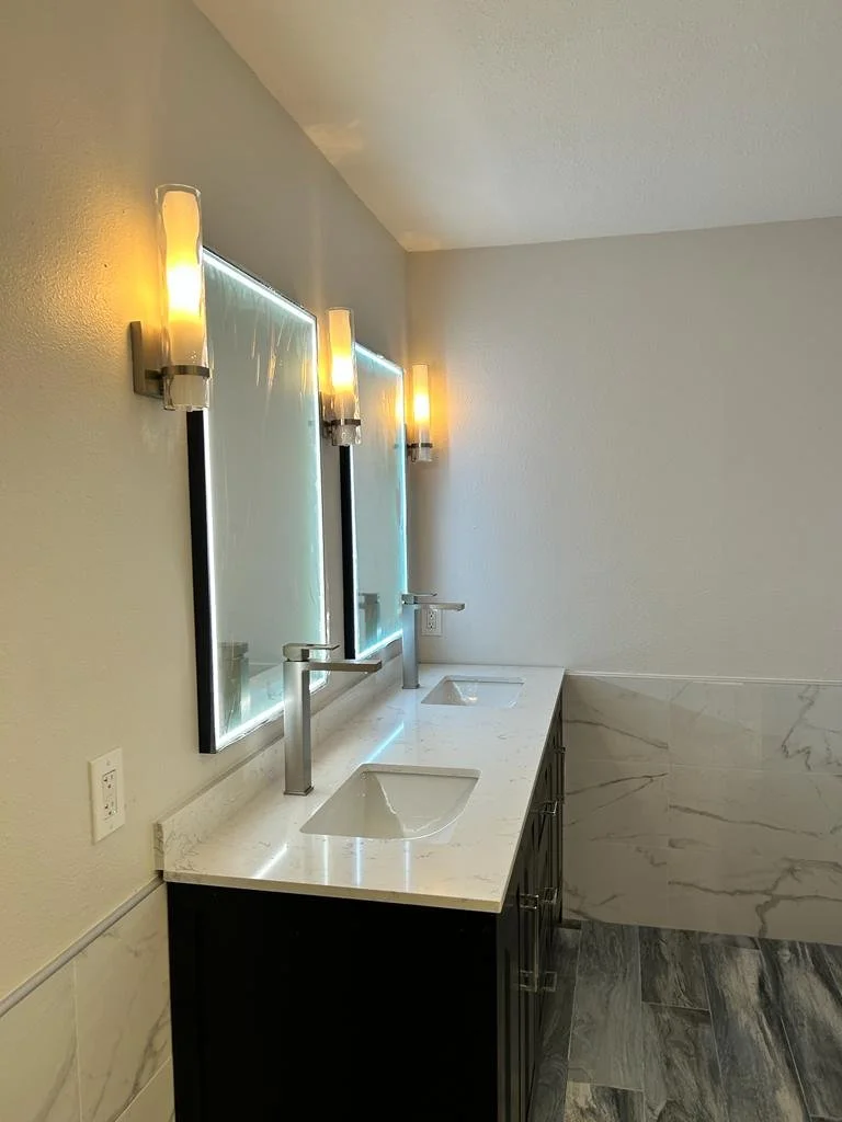 Bathroom remodeling Double bathroom vanity with two mirrors, two modern sinks, and three wall-mounted light fixtures on a white wall with marble tiles on the lower half.