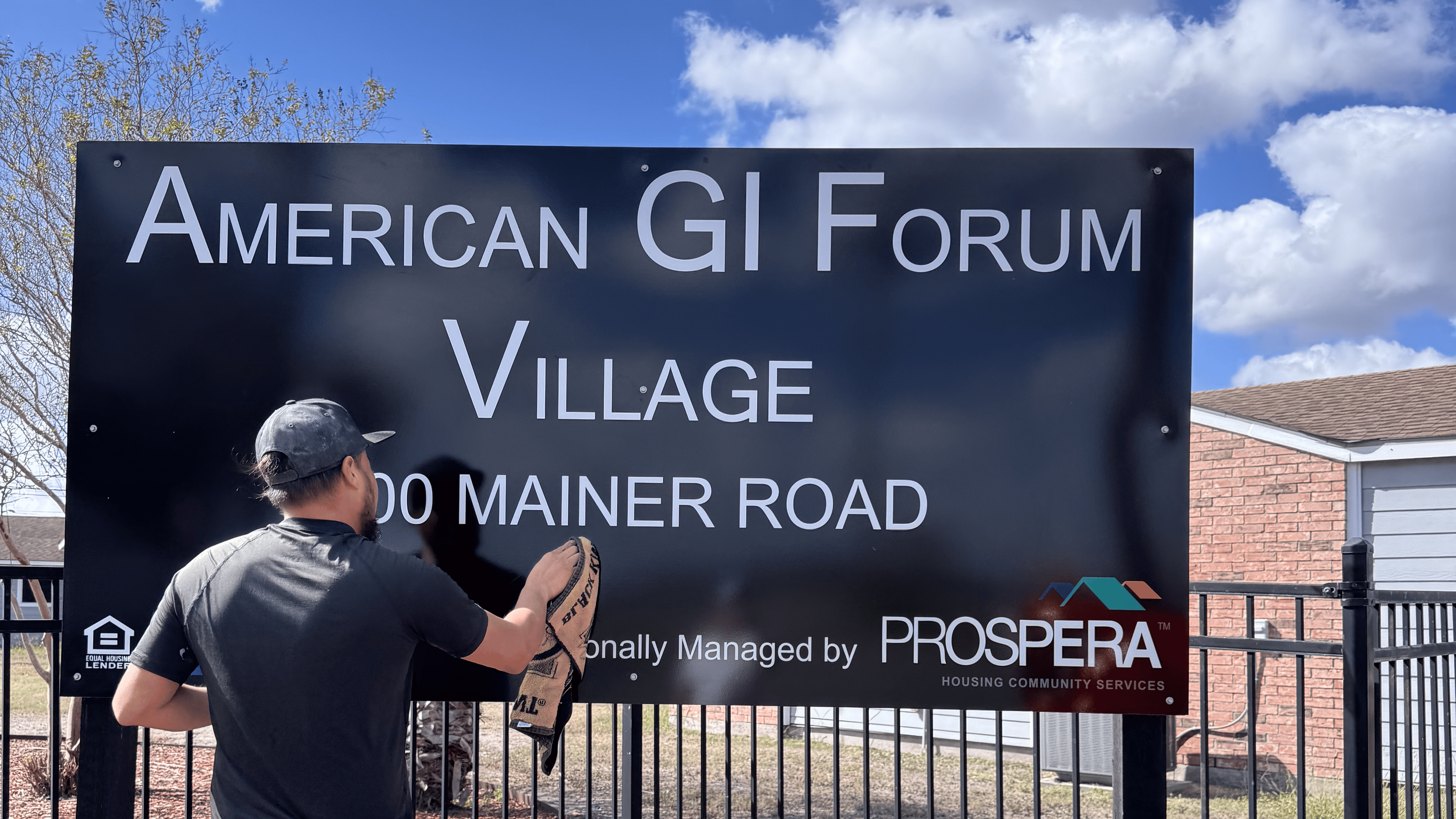 A man cleaning a large black sign with white and red text that reads 'American GI Forum Village, 100 Mainer Road,' with a partly cloudy blue sky and houses in the background.
