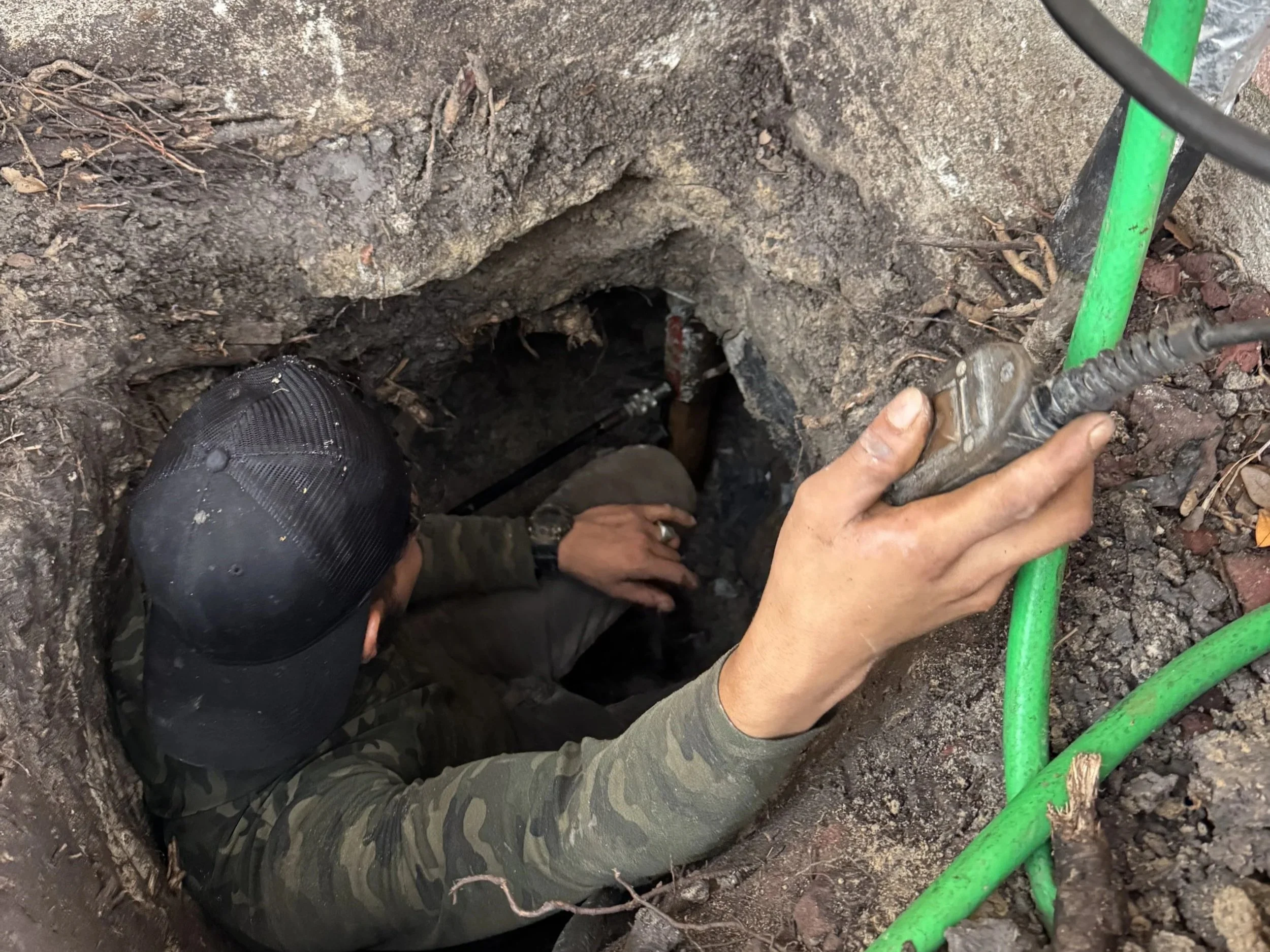 Person in camouflage clothing and black cap working in a ground hole, holding a power tool, with soil and green pipes around the hole.