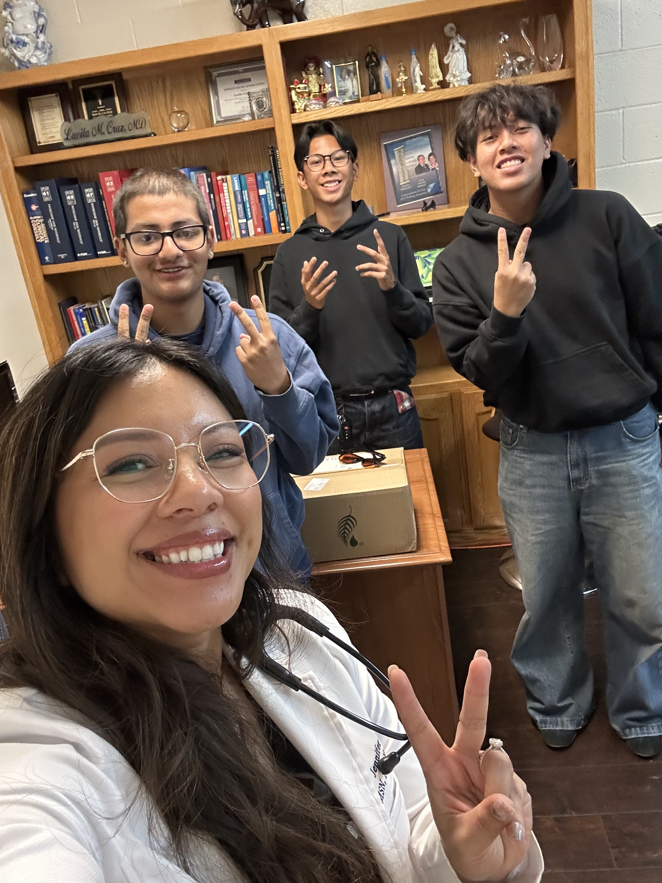 Group selfie of four smiling people making peace signs in front of a bookshelf filled with books, awards, framed photos, and figurines.