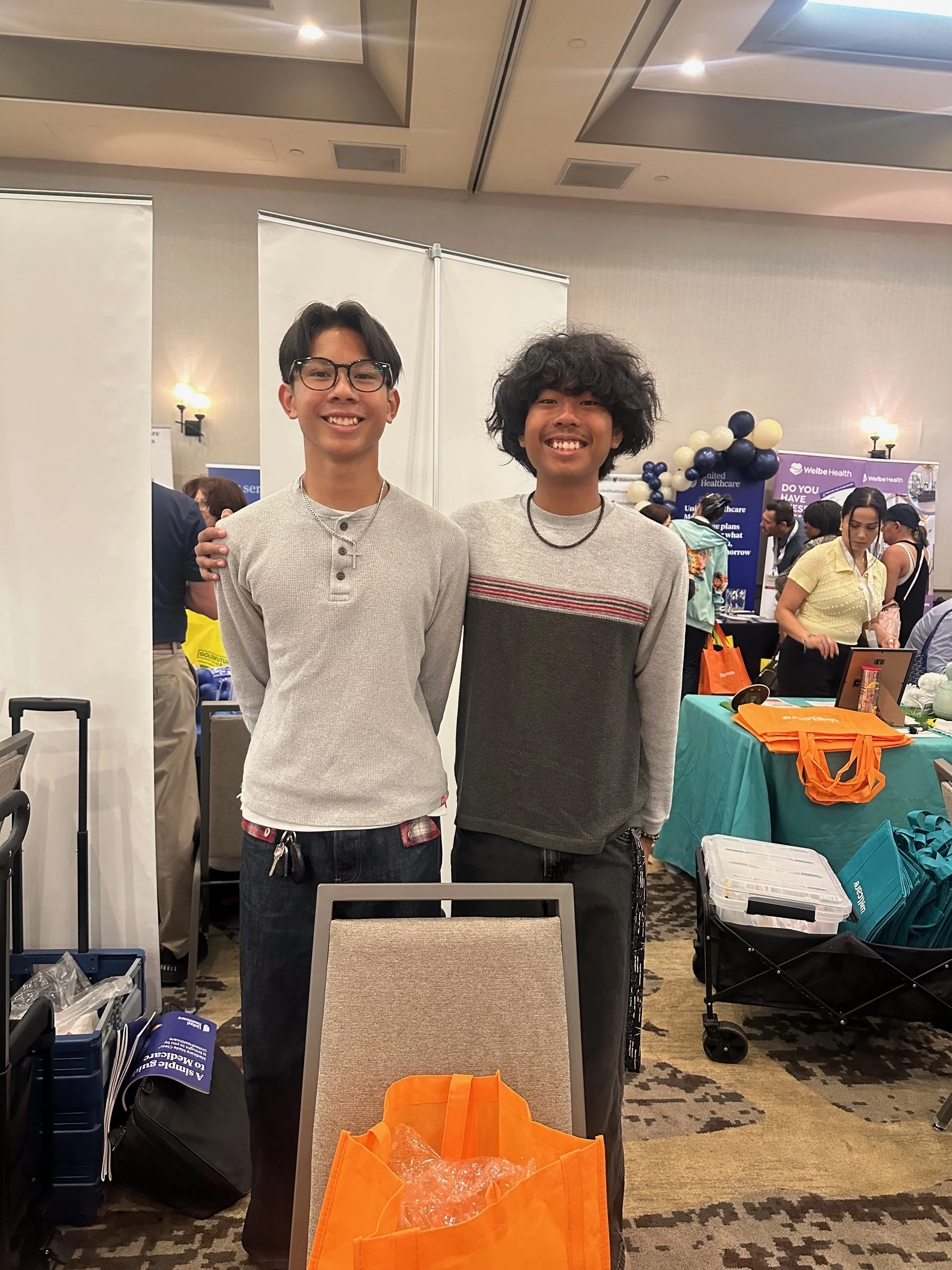 Two smiling young men pose at a healthcare event with vendor booths, balloon decorations, and orange tote bags.