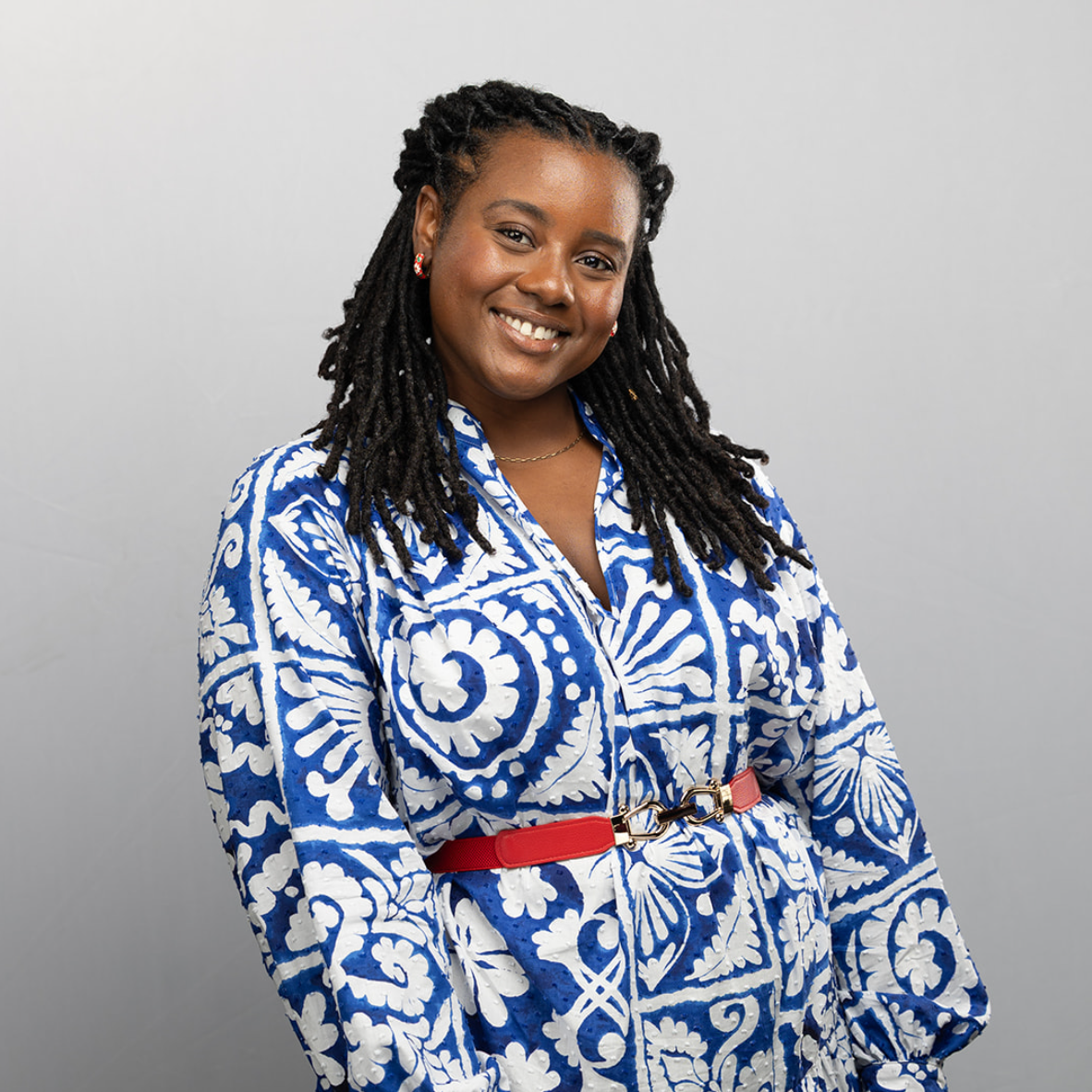Portrait of a smiling Black woman with dreadlocks wearing a blue and white patterned dress with a red belt, standing against a plain gray background.