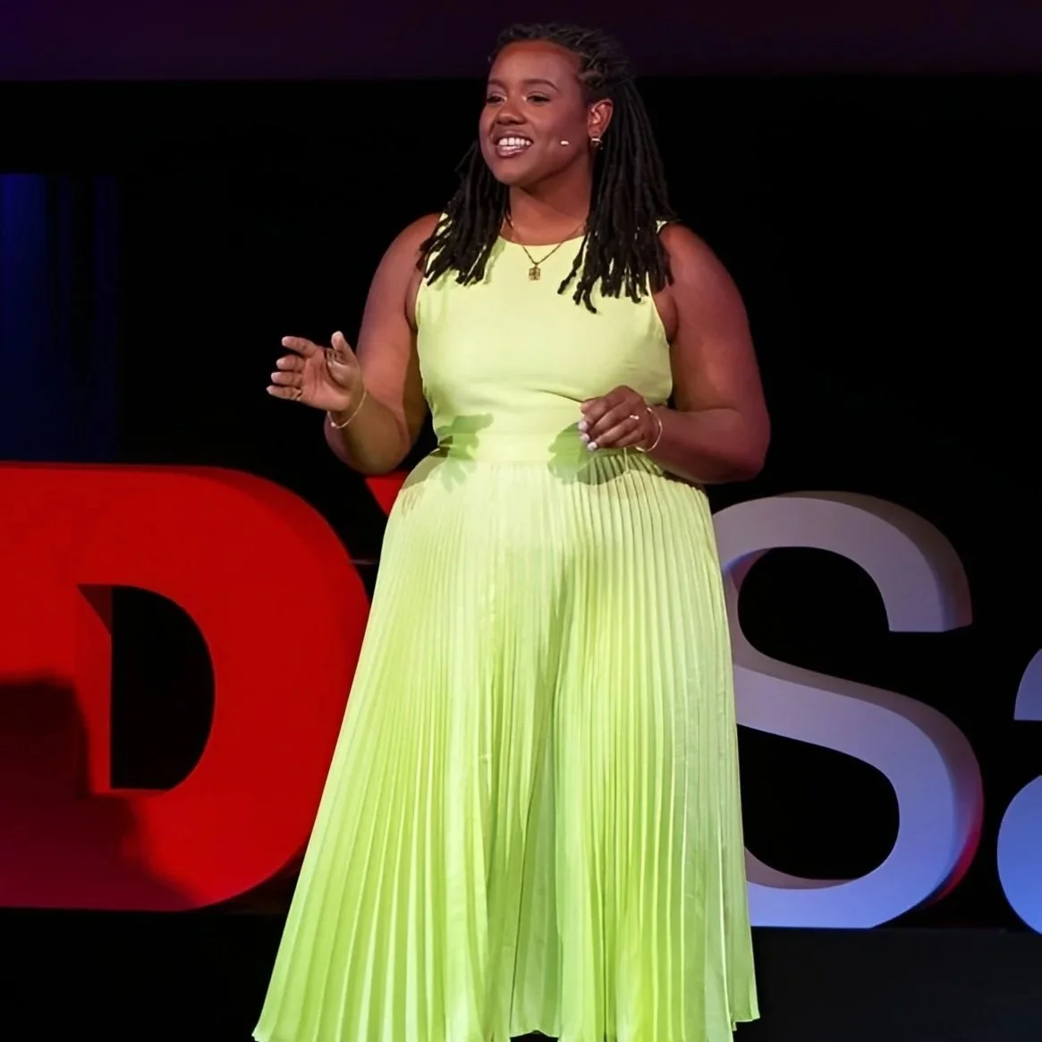 A woman in a bright yellow pleated dress is giving a TED Talk on stage, with a red and black TED logo in the background.