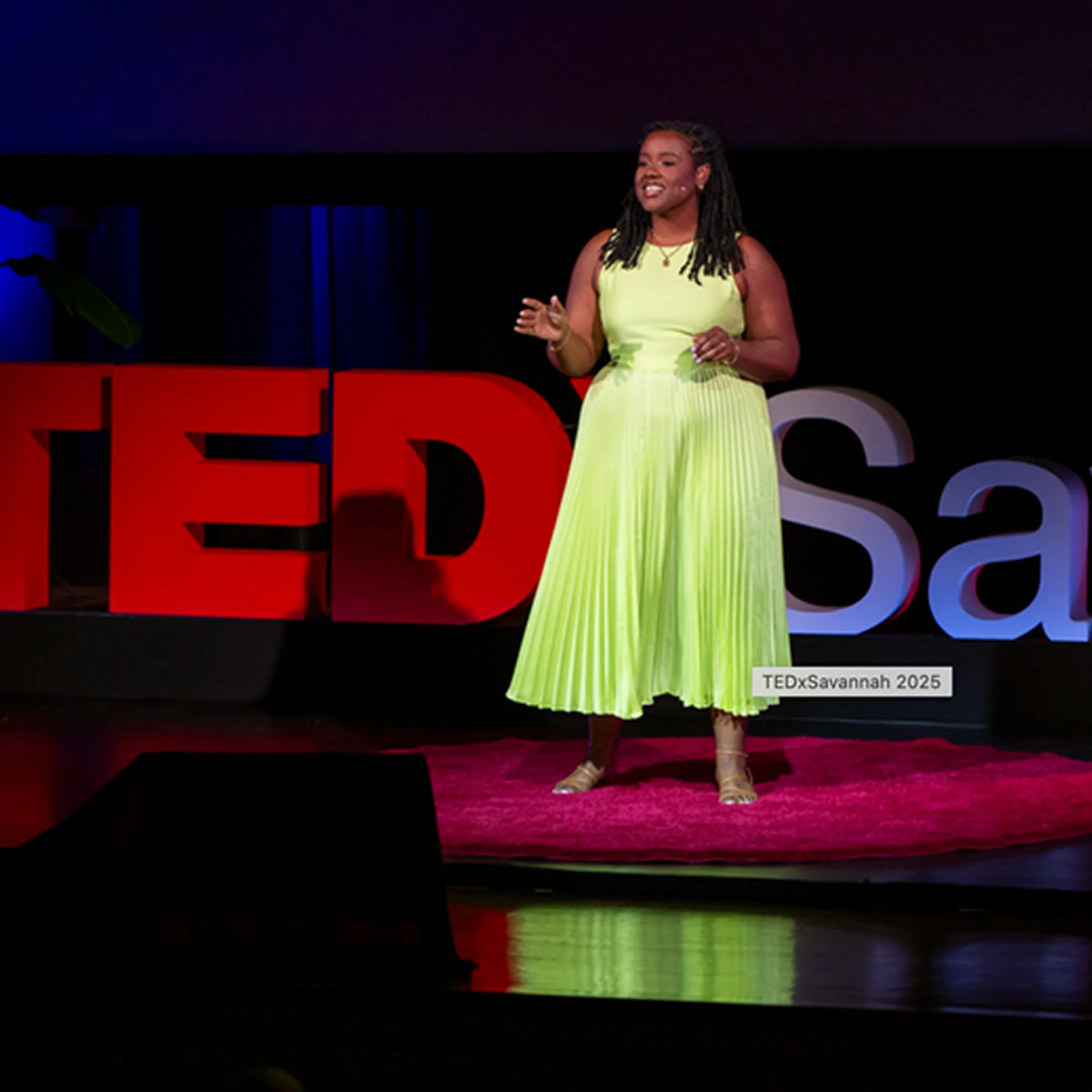 A woman in a lime green dress speaking at a TEDx event on a stage with large red and white TEDx letters, labeled TEDxSavannah 2025.