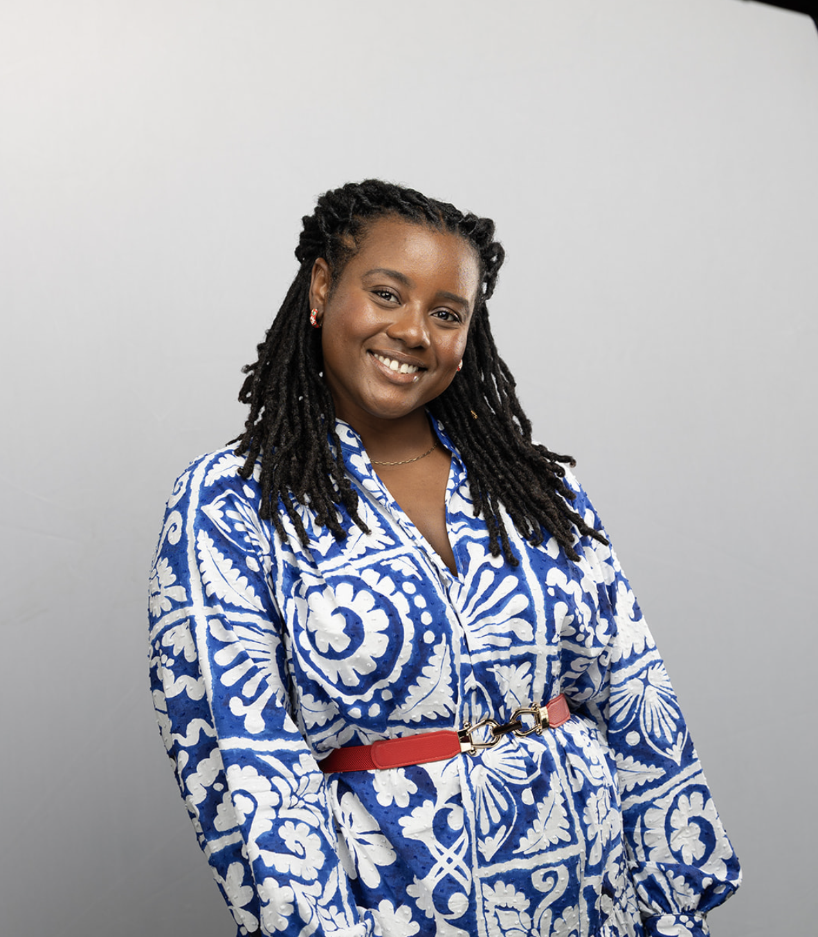 Portrait of a smiling woman with long braids, wearing a blue and white patterned dress and a red belt, standing against a plain gray background.