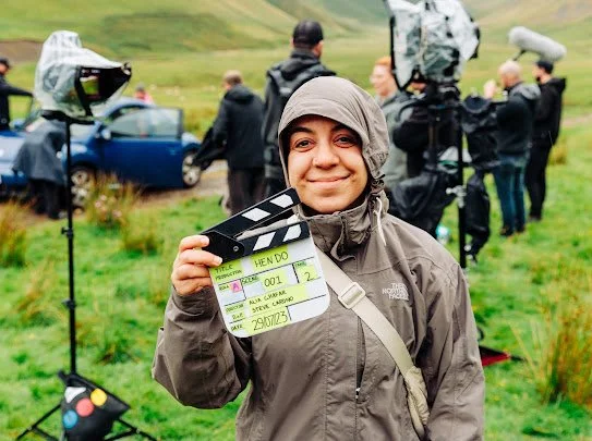 Woman on film set outdoors with crew and equipment in the background, holding a clapperboard.
