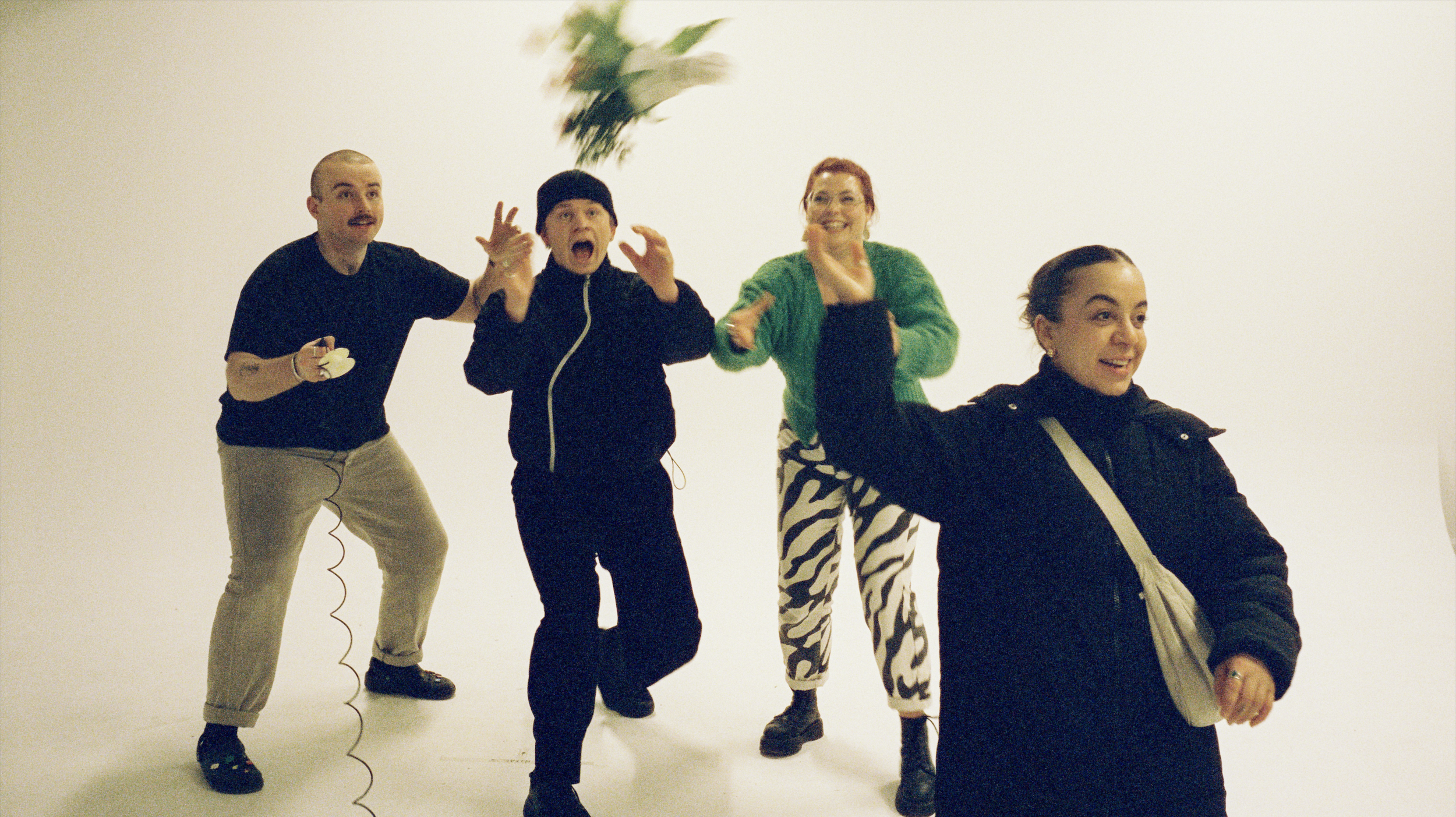 Four people standing against a white background, one throwing flowers in the air, the others reacting with surprise or laughter.