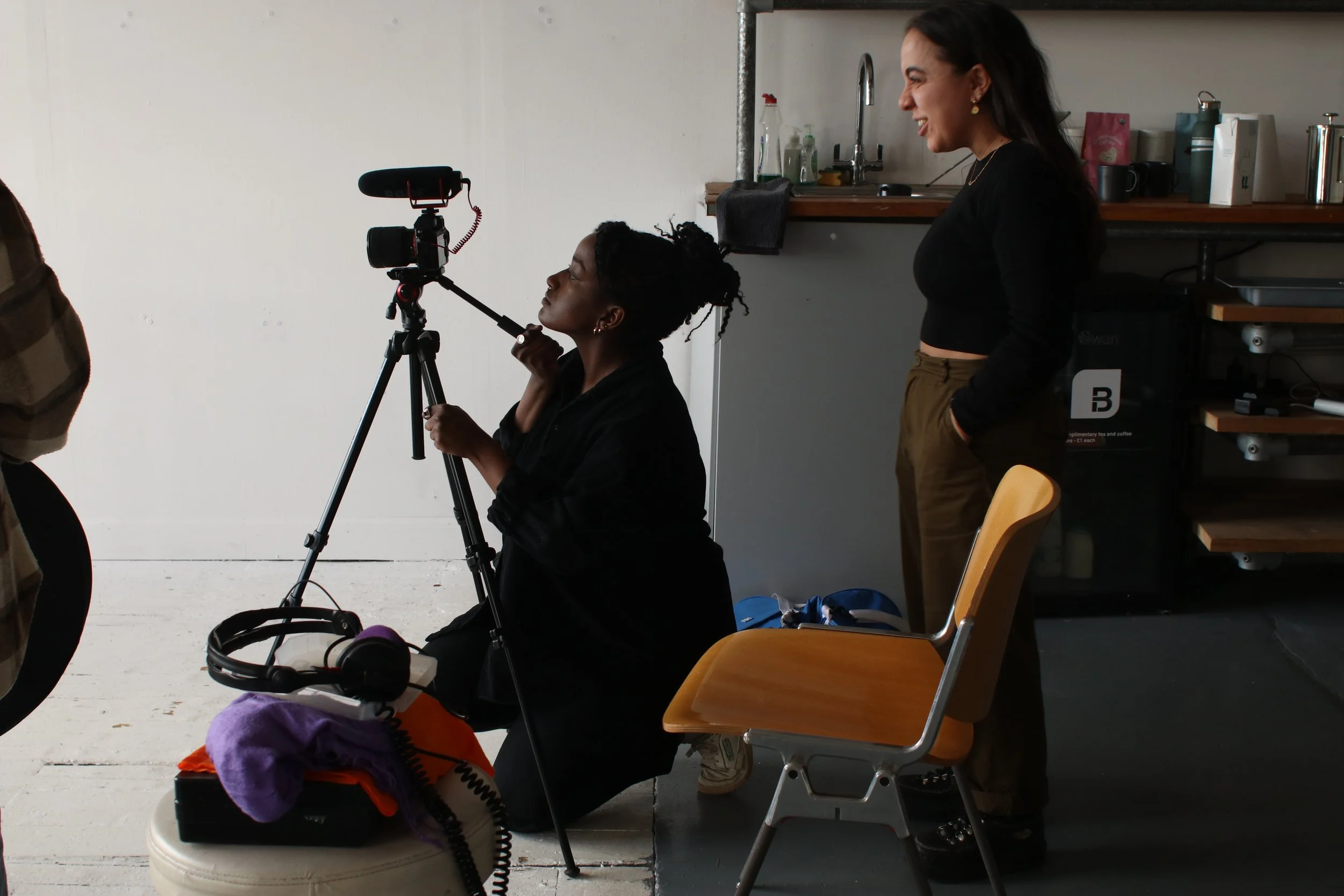 Two women working with filming equipment in a studio. One woman is sitting down, operating a camera on a tripod, while the other woman stands nearby, smiling and looking at her contributor.