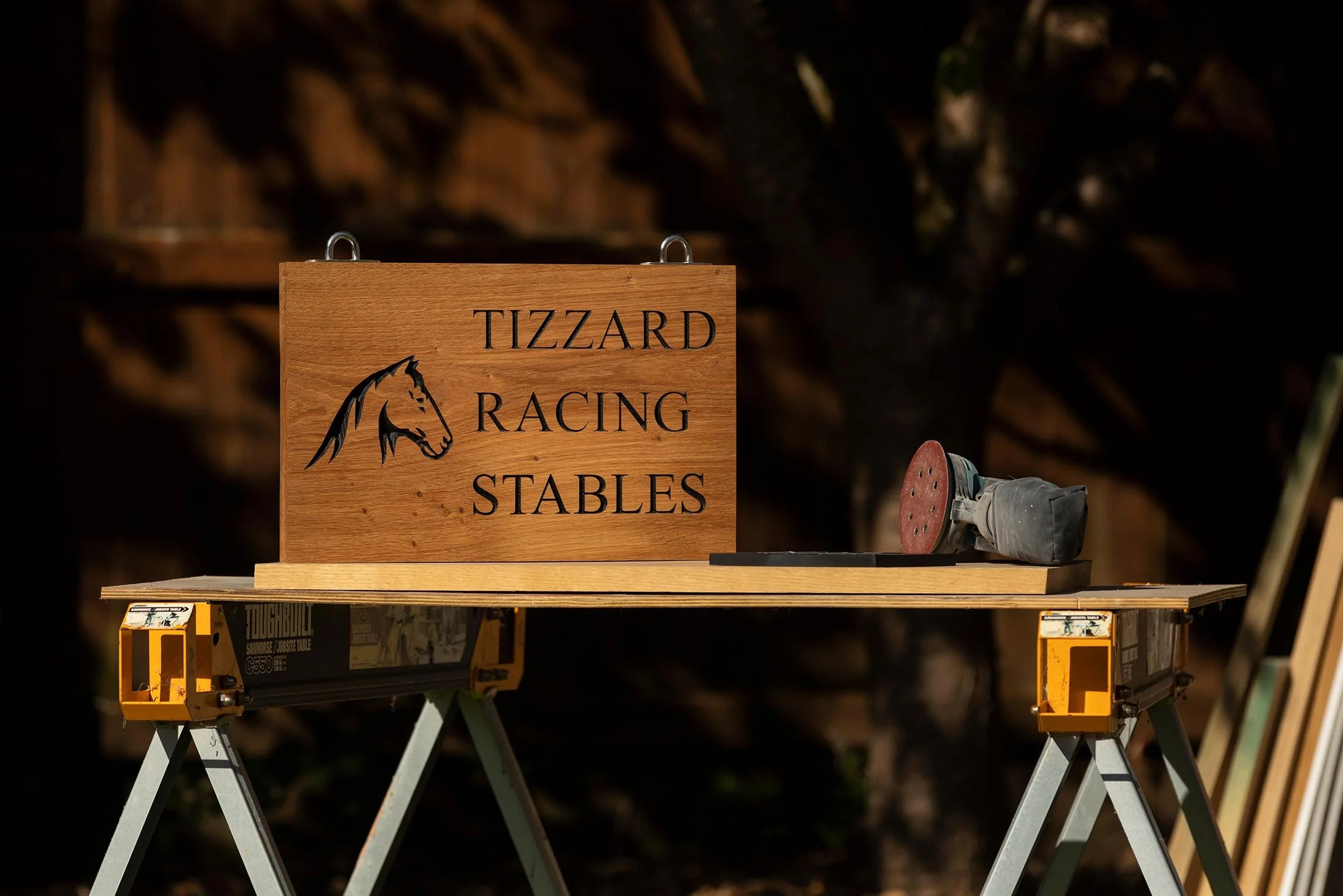 A wooden sign with black text reading 'Tizzard Racing Stables' and a horse head logo. The sign is placed on a workbench with a power sander to the right. The background is dark and blurry.