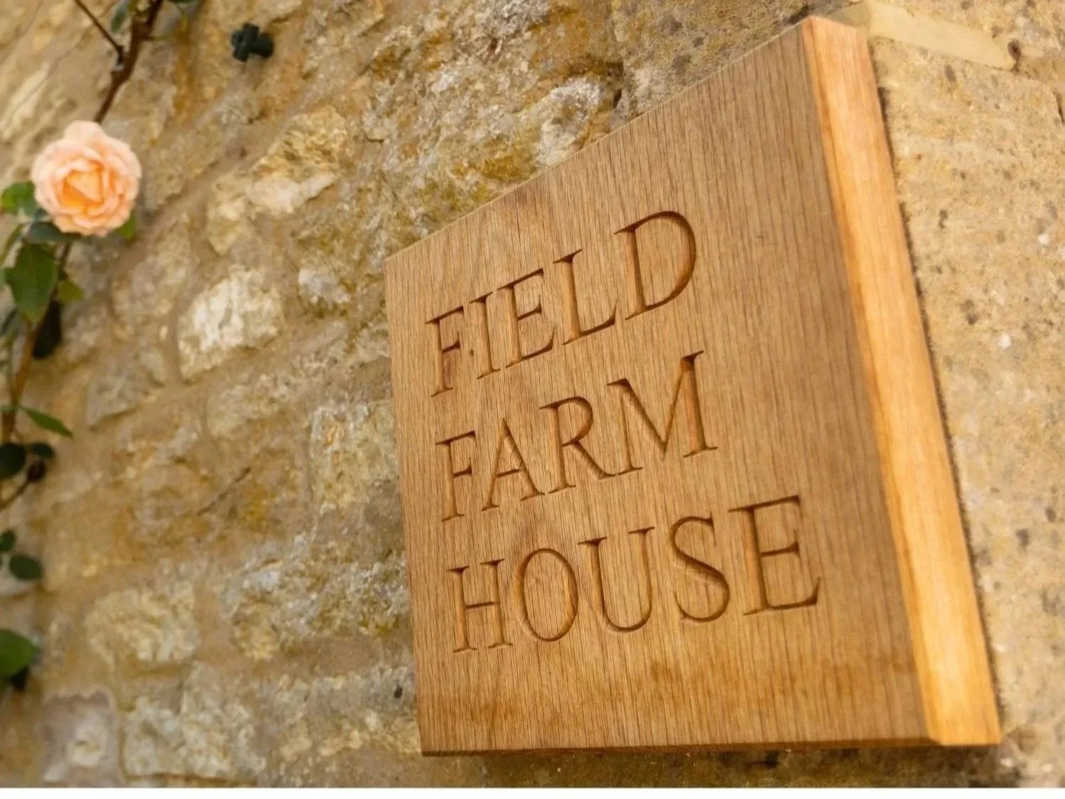 Wooden sign on stone wall that reads 'FIELD FARM HOUSE' with a pink flower partially visible on the left side.