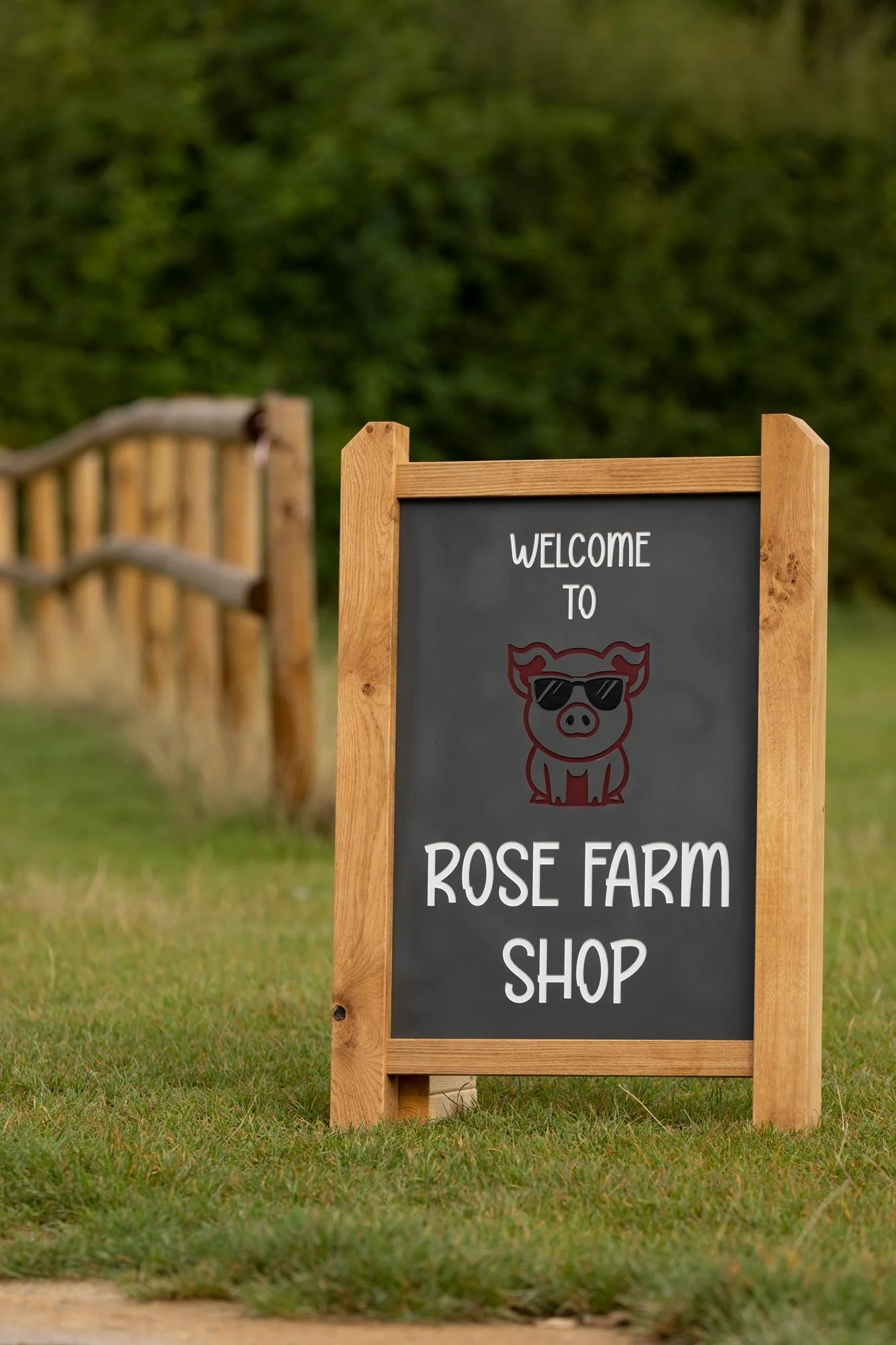 Wooden-framed chalkboard sign reading 'Welcome to Rose Farm Shop' with a pig wearing sunglasses illustration, on a grassy area with a wooden fence and green hedges in the background.