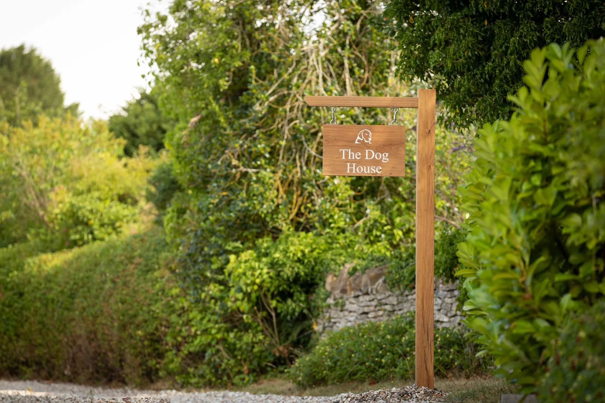 A wooden sign hanging from a post reads 'The Dog House' with a small dog illustration above the text, set against a background of green bushes and trees.