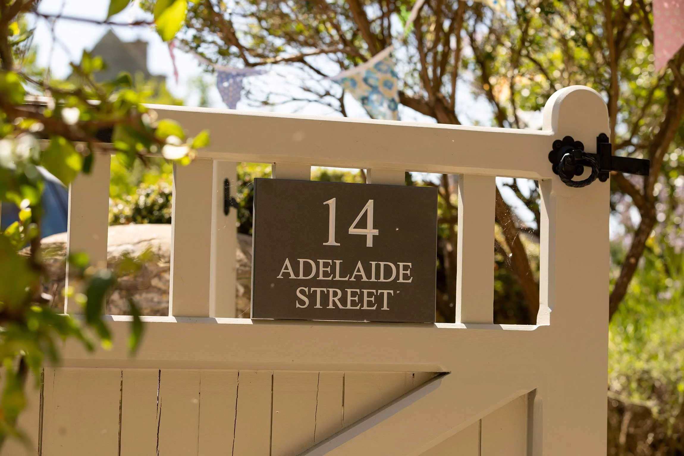 A white picket fence with a black address plaque that reads "14 ADELAIDE STREET". There are colorful bunting flags hanging in the background, with leafy trees and a clear sky.