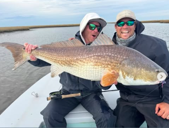 Two men in sunglasses and hats on a boat holding a large redfish.