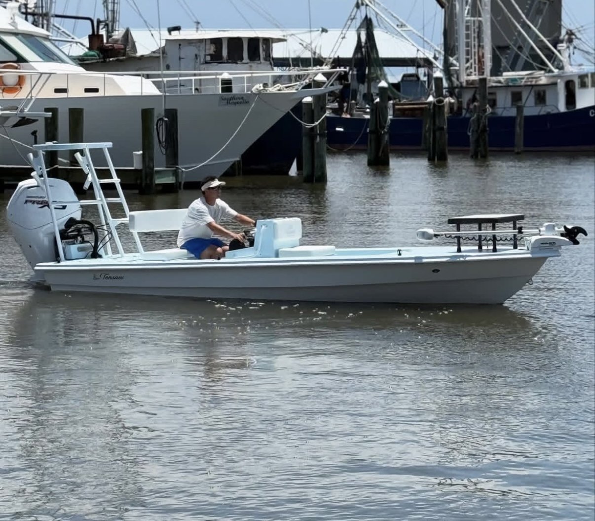 A man in a white shirt, blue shorts, and a hat steering a small white motorboat near a marina with larger boats docked in the background.