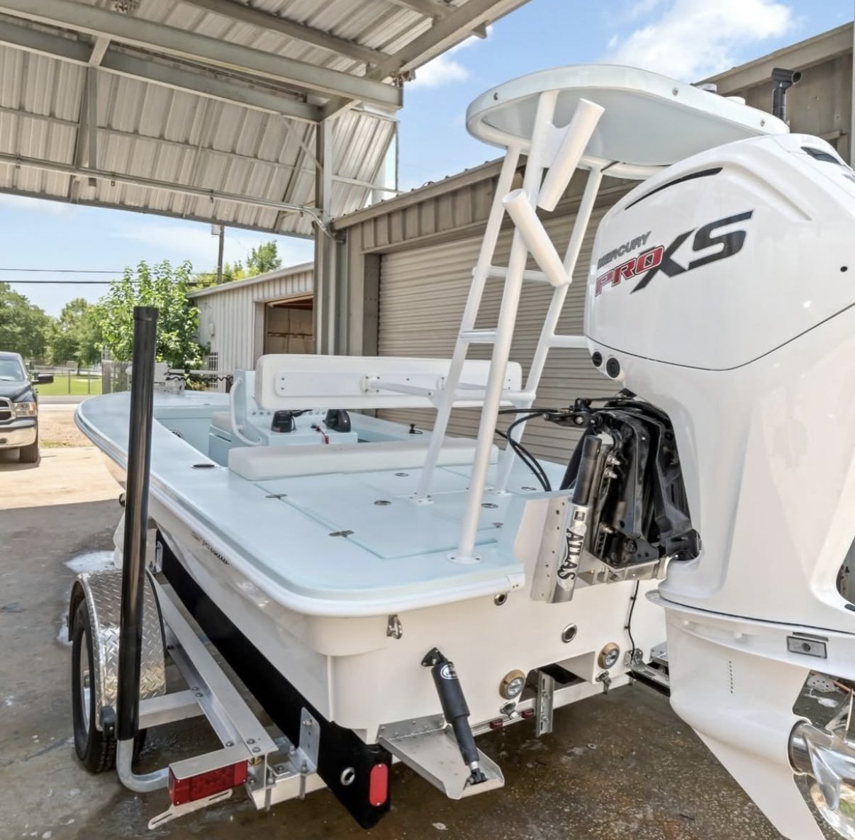 A white boat with a Mercury Pro XS outboard motor on a trailer, parked outside near a building with a metal roof and a garage door.
