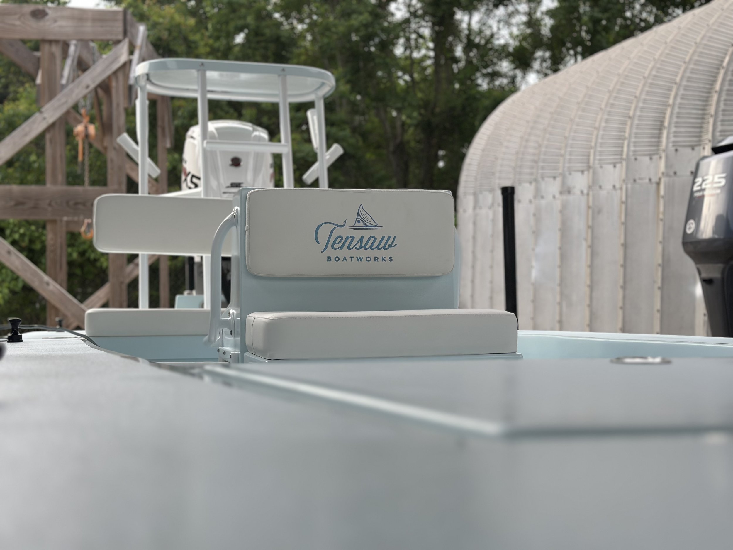Close-up of a light blue Tensas Boatworks boat with a padded seat back branded with the company logo, with a wooden deck and a garage in the background.