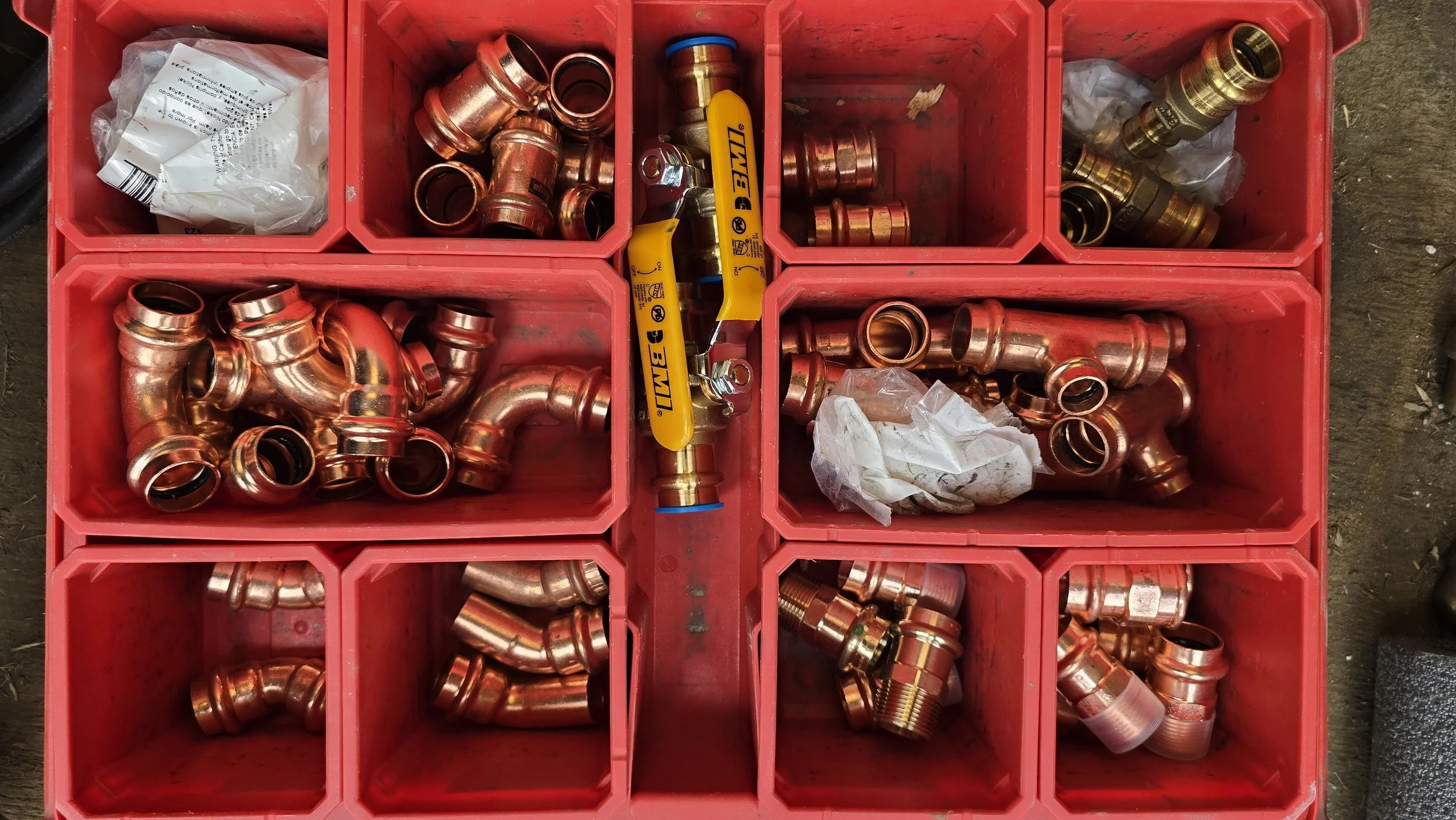 Red plastic container divided into compartments holding various copper plumbing fittings, including elbows, couplings, and adapters, with yellow and blue-handled tools in the center compartments.