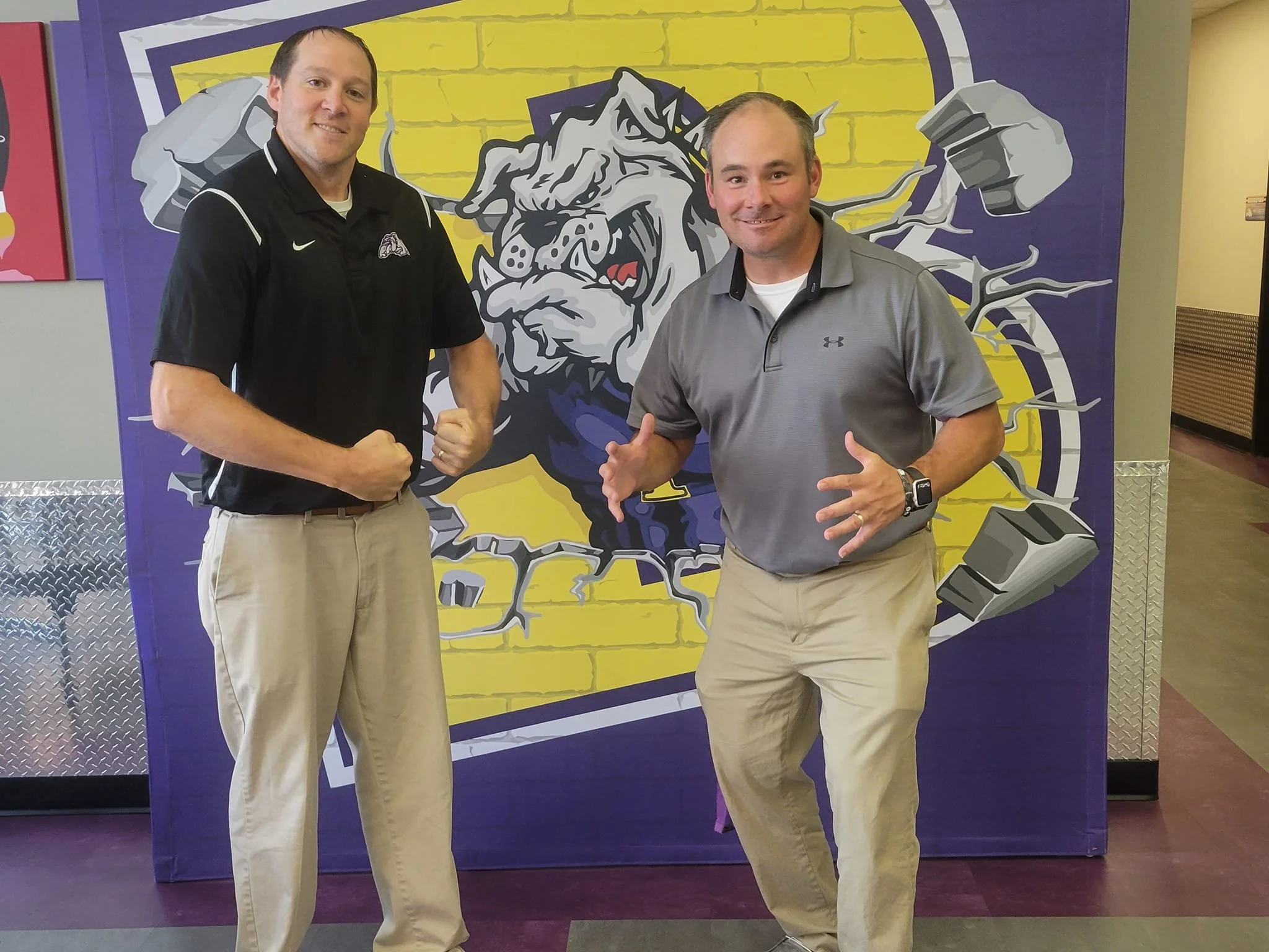 Two men standing indoors in front of a large, colorful mascot mural. One man is wearing a black polo shirt and khakis, and the other man is wearing a gray polo shirt and khakis. Both are posing with fists and hands raised, smiling at the camera.