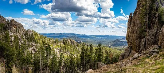 A panoramic view of a mountainous landscape with rocky cliffs, dense green trees, and a partly cloudy sky.