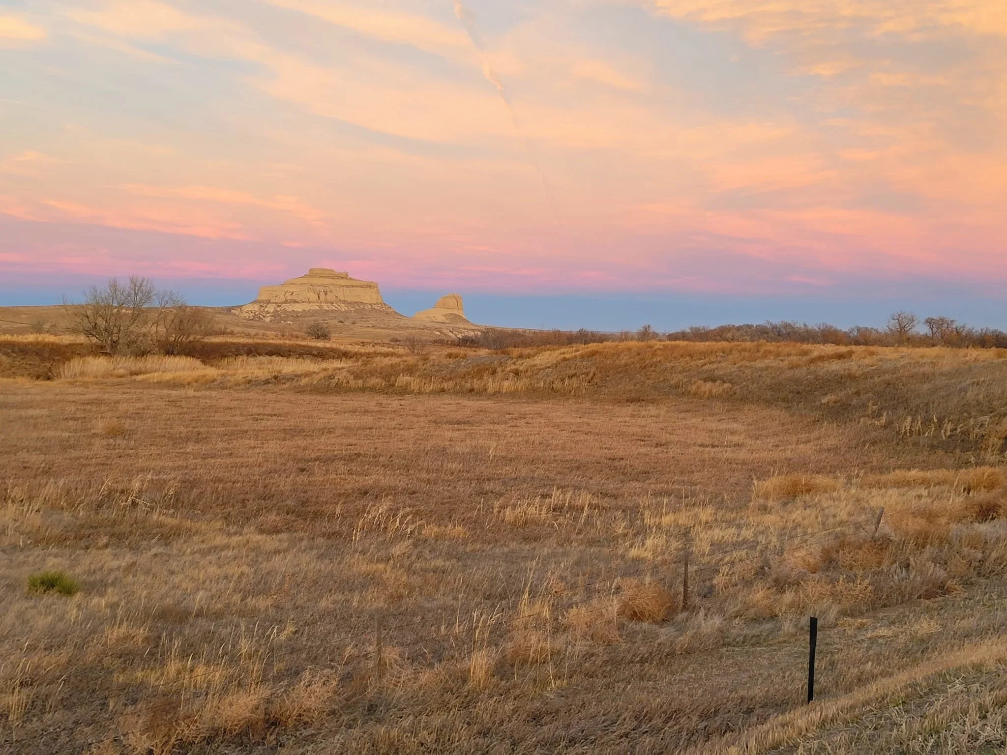 A scenic landscape of a grassy field with a distant rock formation under a pink and blue sky at sunset.