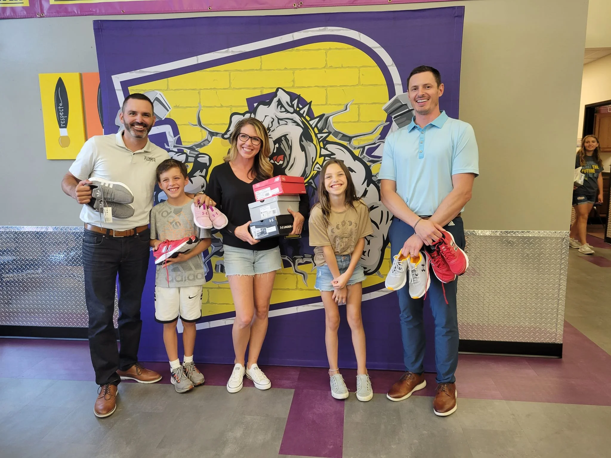 Group photo of three adults and two children standing indoors in front of a wall mural of a bulldog mascot. The adults are holding sneakers, and one child is holding a pair of shoes. Everyone is smiling.