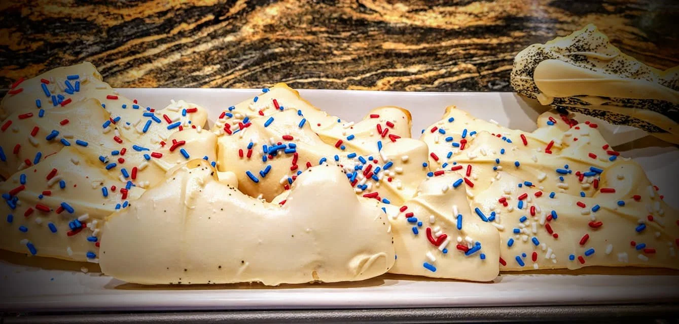 A white rectangular plate with a decorated cream-filled pastry topped with red, white, and blue sprinkles, placed on a wooden surface.