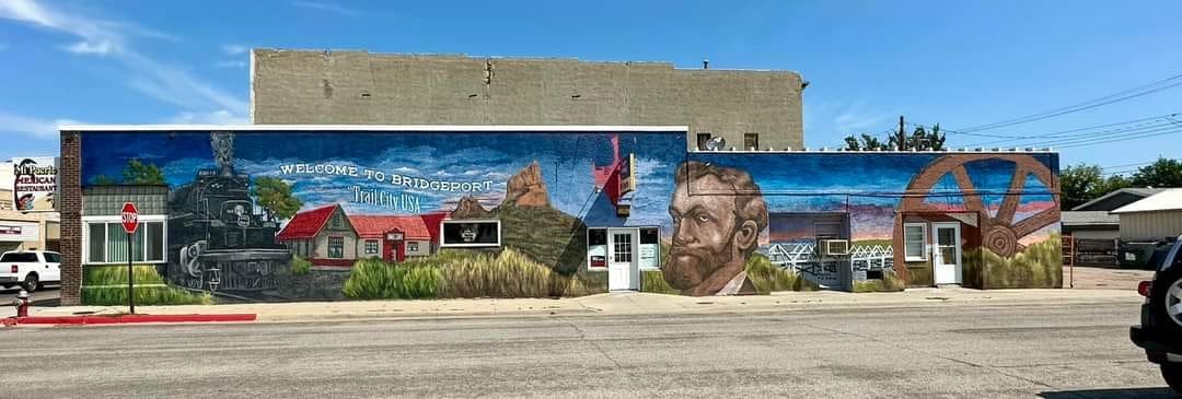 Colorful mural on building wall in Bridgeport, USA, featuring a train, historic buildings, a mountain landscape, a portrait of a man with a beard, and large wheels or gears in the background.