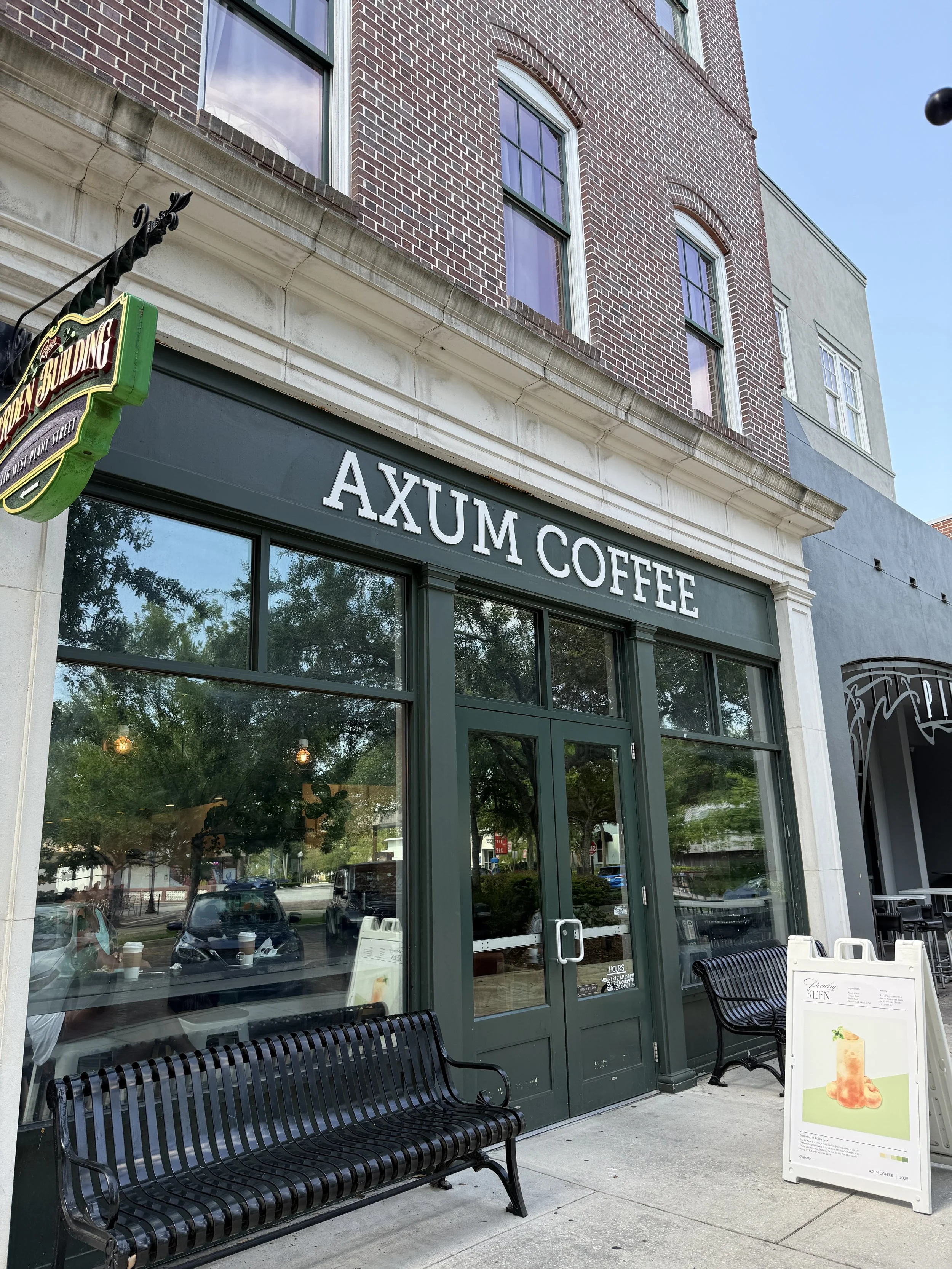 Exterior of Axum Coffee shop with large glass windows, black bench outside, sidewalk signboard, and brick building facade with upper window views.