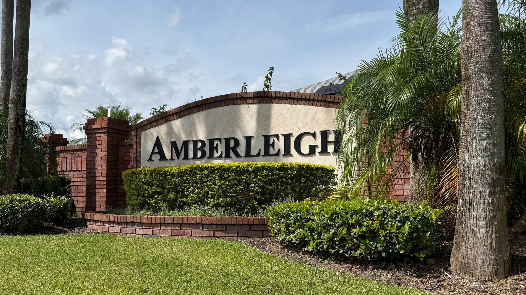 Community entrance sign for Amberleigh in Winter Garden, Florida. Brick and stucco monument with black raised lettering, surrounded by manicured hedges, palm trees, and green lawn under a partly cloudy sky.