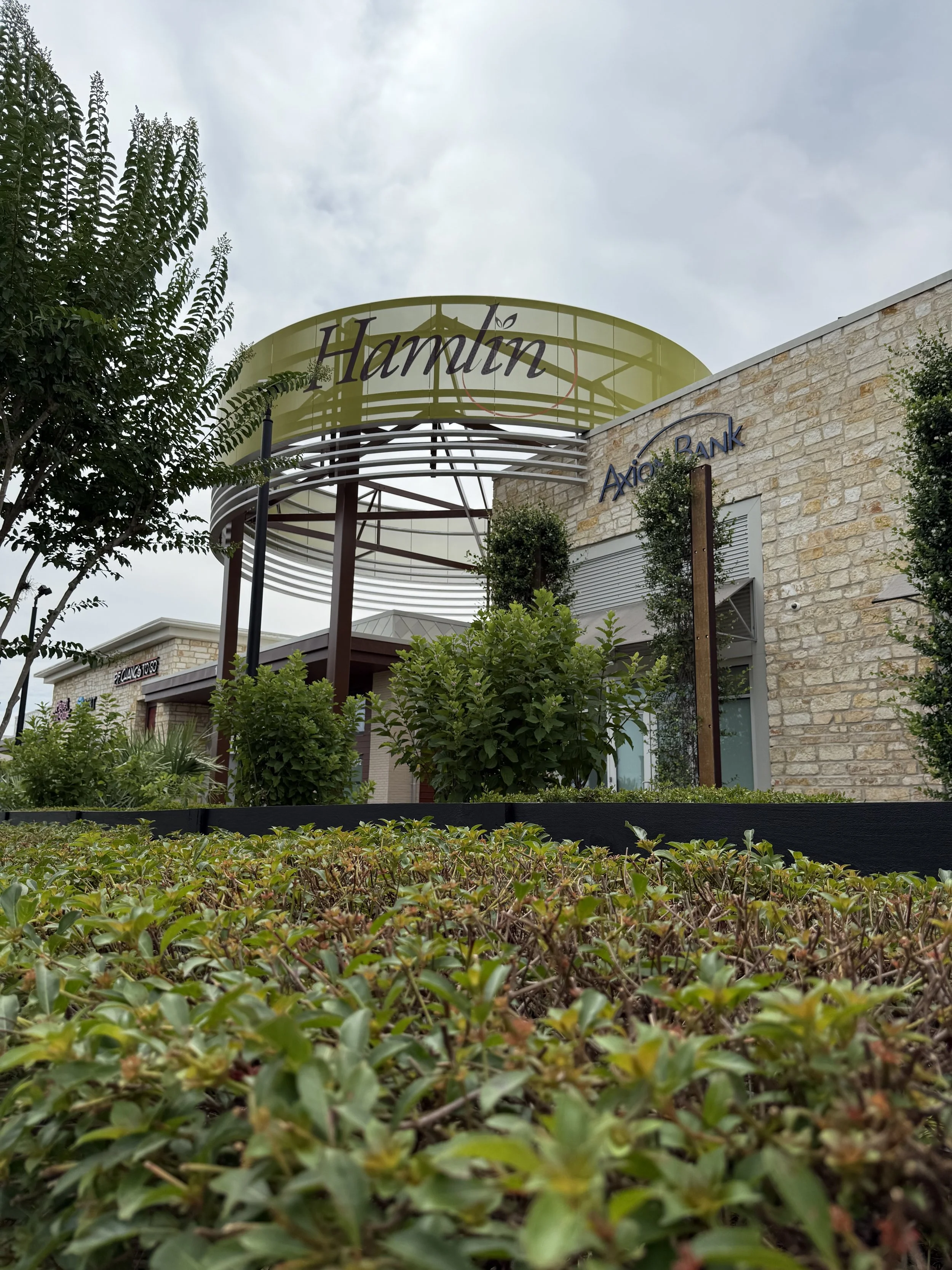 View of a commercial shopping center entrance with plants and bushes in the foreground, a large sign that says 'Hamlin,' and a stone building with a sign that says 'Axis Bank' on it.