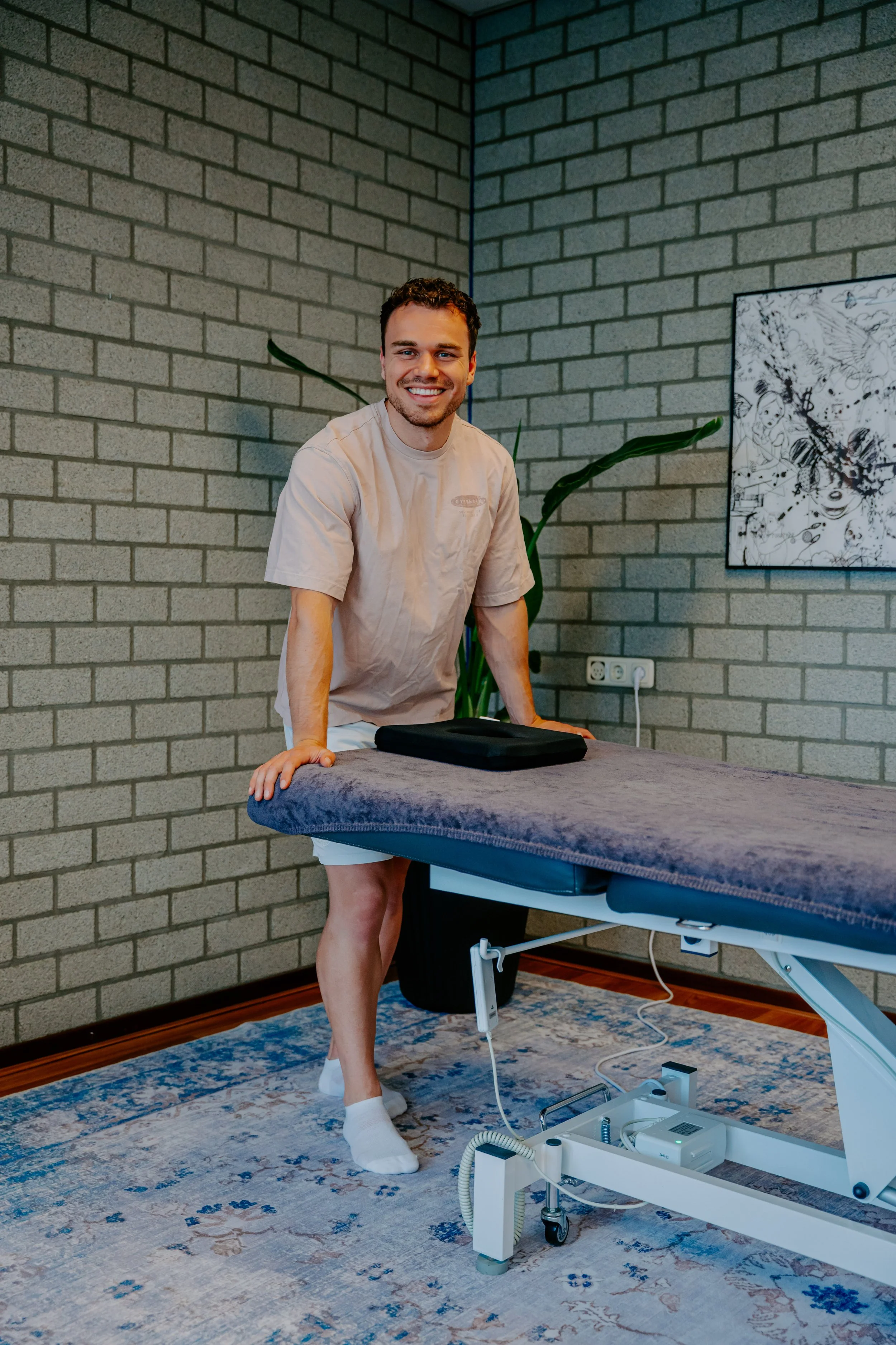 Een man in slaaptextiel staat naast een massage tafel die bedekt is met een blauw bedekking. De man glimlacht en kijkt naar de camera. Op de achtergrond een muur van grijze bakstenen, een grote plant en een zwart-wit kunstwerk aan de muur. De vloer is bedekt met een blauw en beige tapijt. Een elektrische massagetafel met bedieningspanelen en een kabel ligt op de vloer.