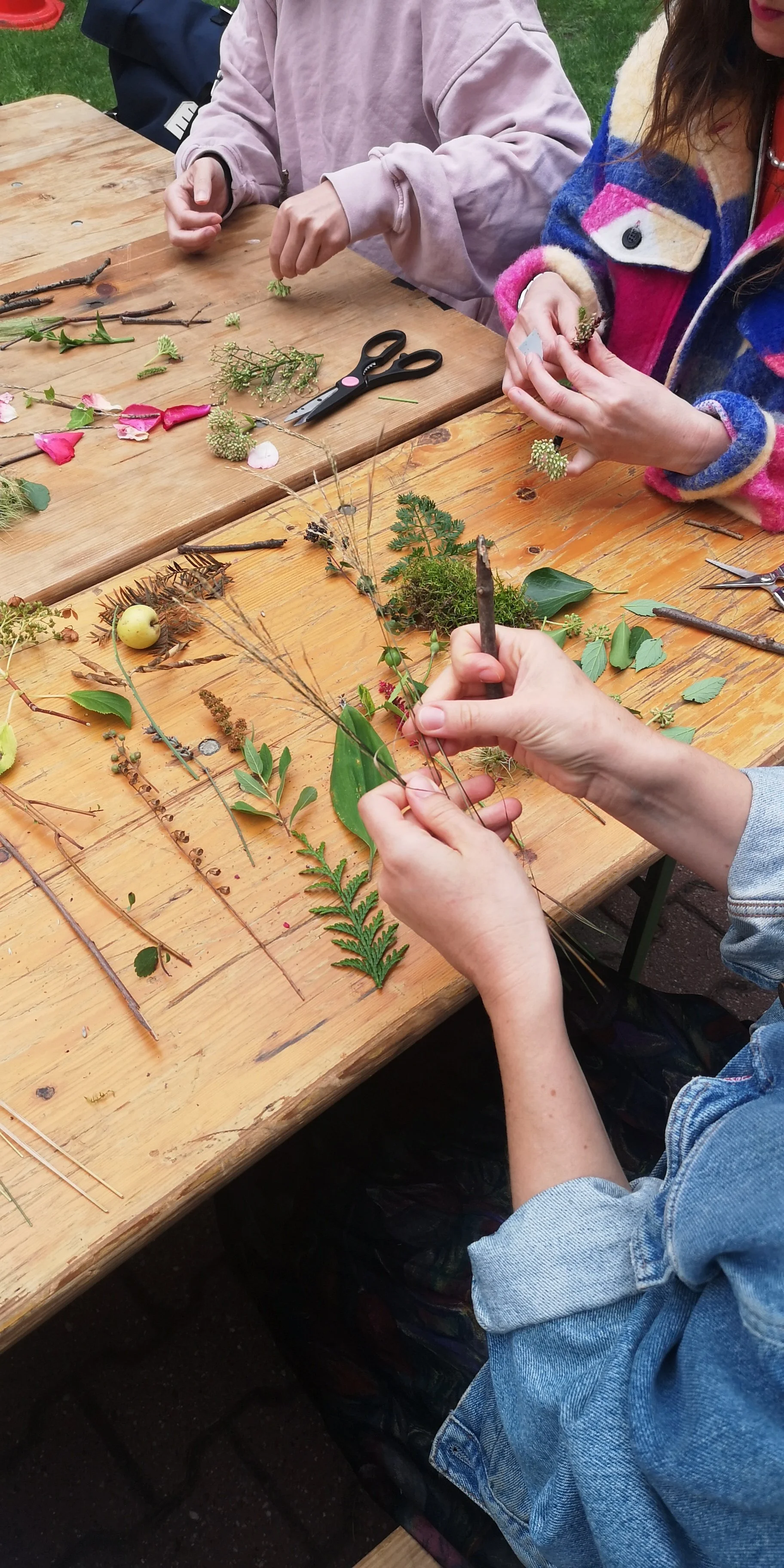 Persone che creano un collage di elementi naturali come foglie, ramoscelli e fiori su un tavolo di legno durante un’attività all’aperto.