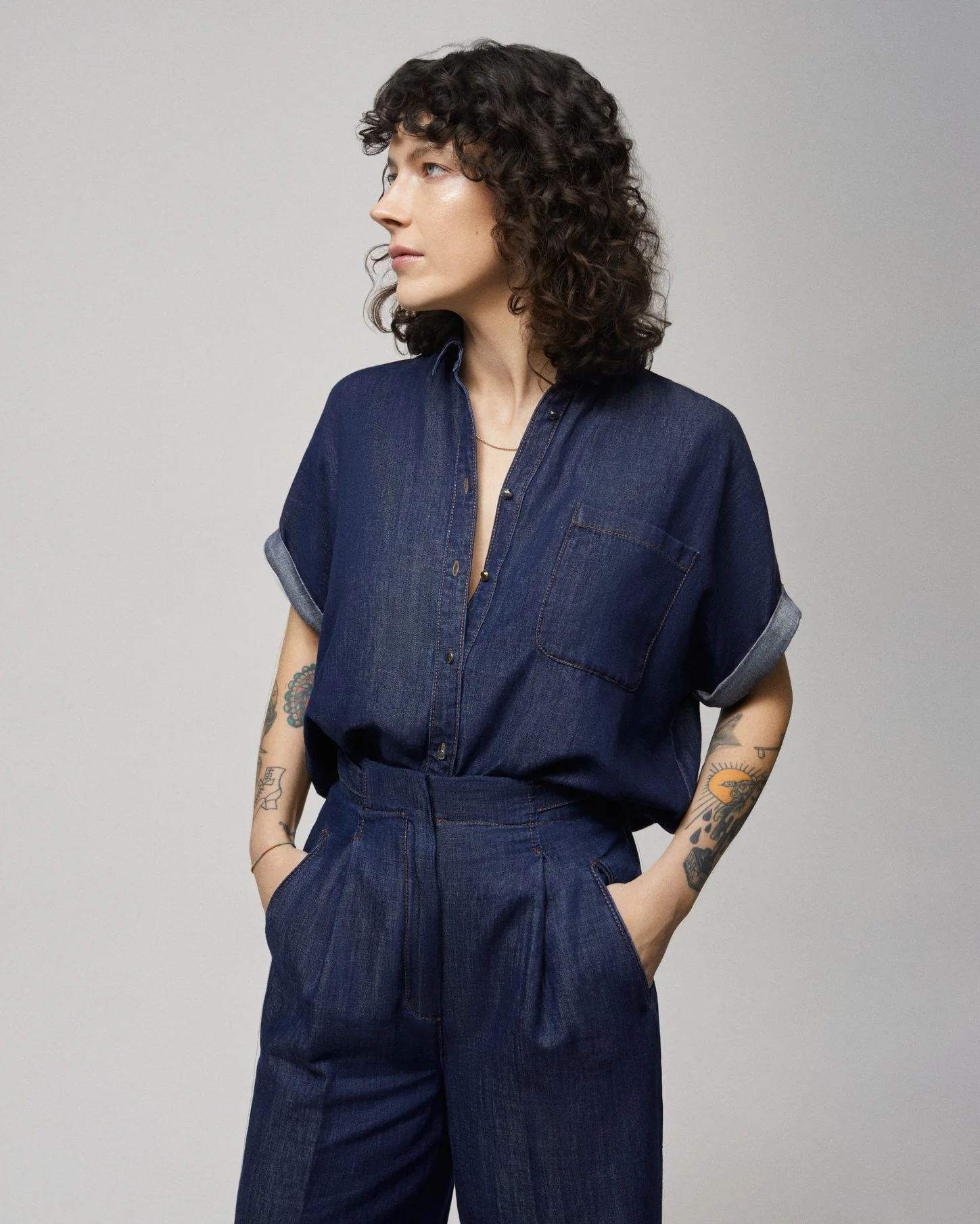Una donna con capelli riccioli corti in posa contro uno sfondo grigio, indossa un completo in denim blu con camicia a maniche corte e pantaloni.