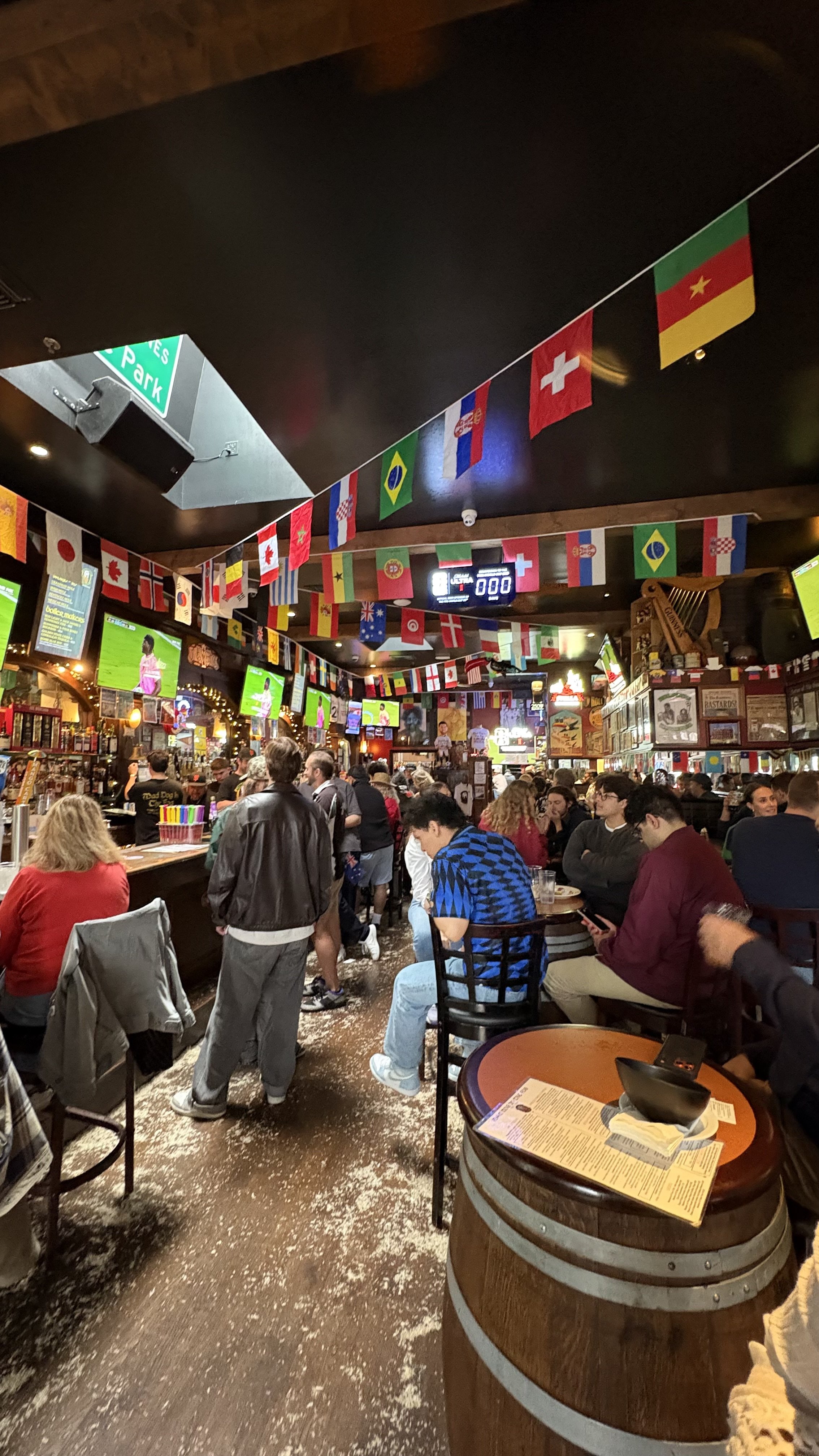 A crowded bar or restaurant decorated with international flags hanging from the ceiling, with patrons sitting and standing while engaging in conversations, watching screens, and ordering drinks.