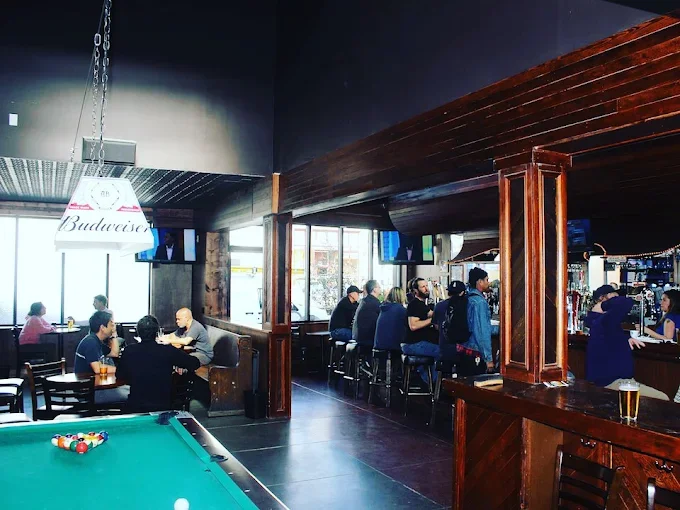 People sitting and socializing at a bar with a pool table in the foreground, large windows letting in natural light, TVs on the walls, and wooden accents inside the bar.